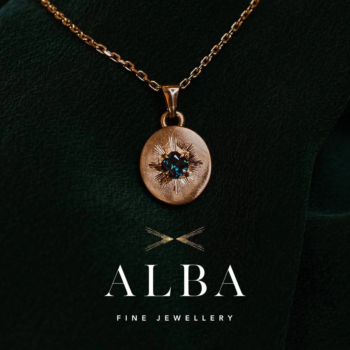 Gold pendant necklace with a dark gemstone, featuring a sunburst design, on a person wearing a black top at ALBA Fine Jewellery.