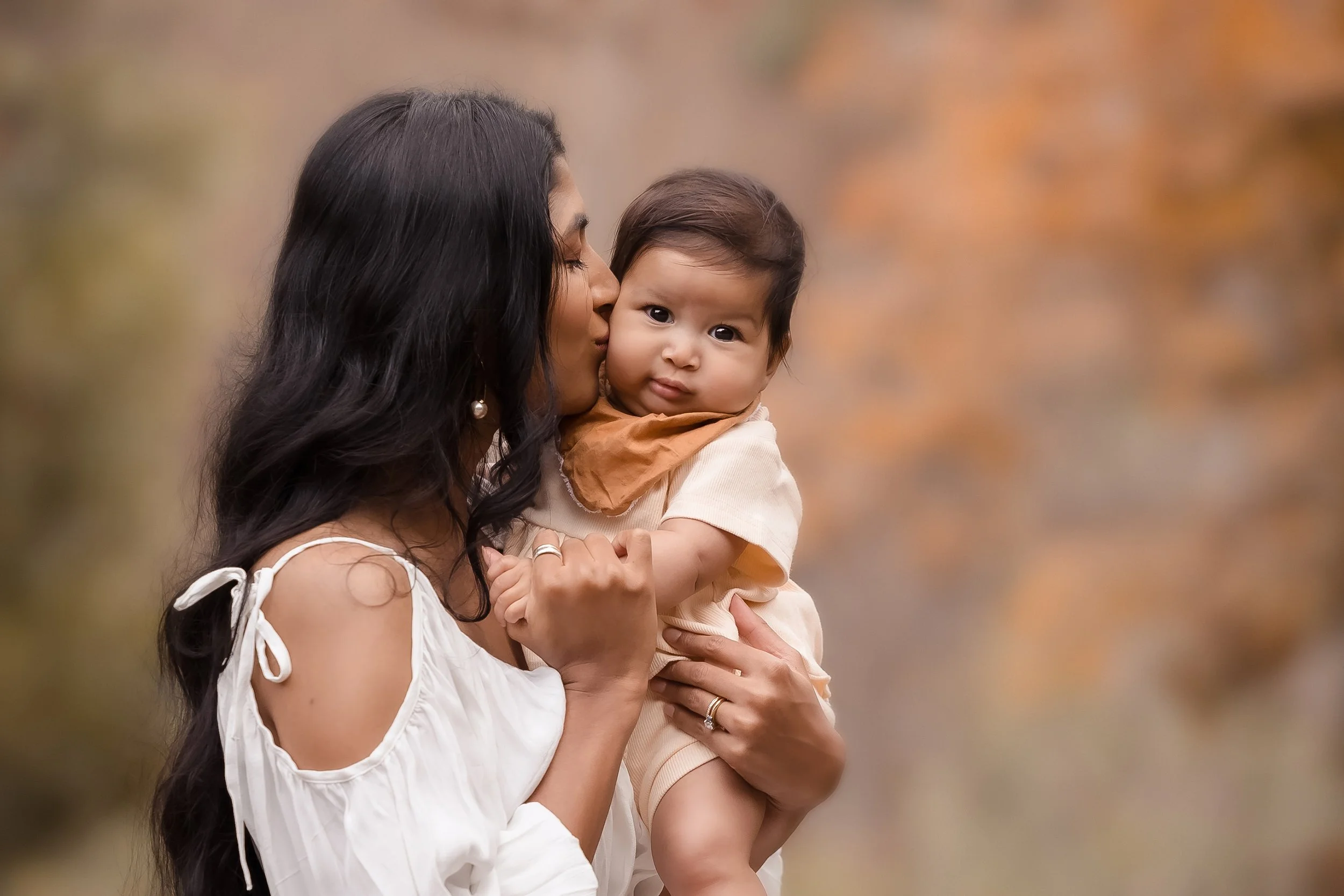 A woman with long black hair holding a young child with dark hair outdoors, with a blurred background of trees and autumn leaves.