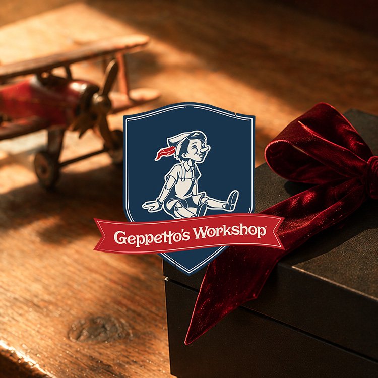 Close-up of a gift box with a large red velvet ribbon, a logo with a cartoon girl sitting on a shield, and the text 'Geppetto's Workshop'; a blurred wooden toy airplane in the background.
