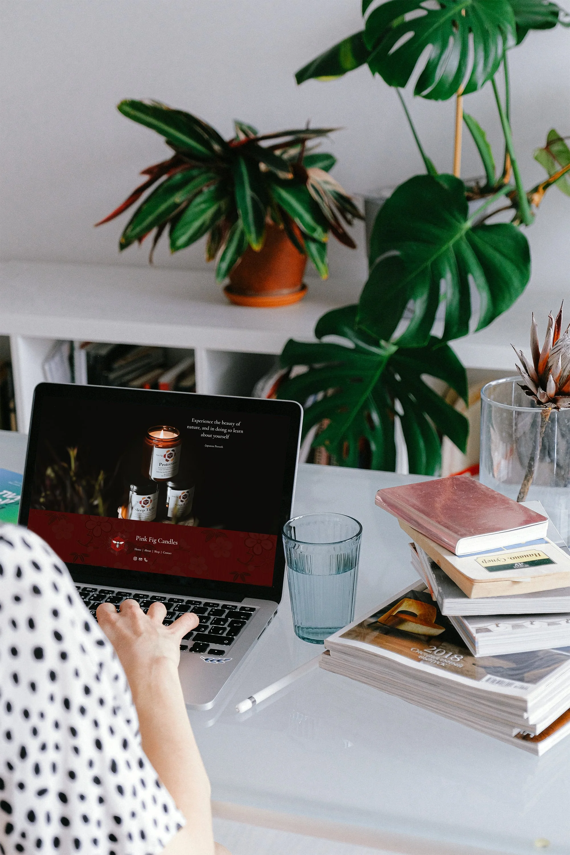 A person typing on a laptop at a white desk with a stack of books, a glass of water, and a plant in a glass container, with large green-leafed plants in the background.