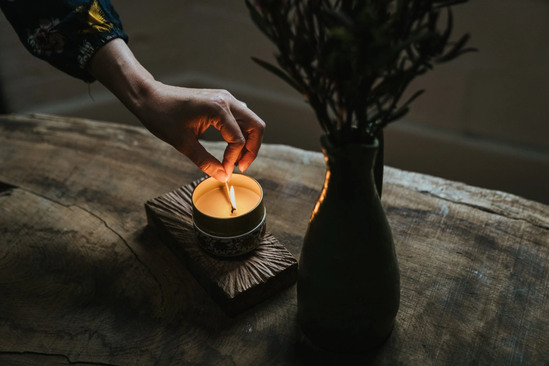 A person lighting a candle on a wooden table next to a large dark vase with a plant.