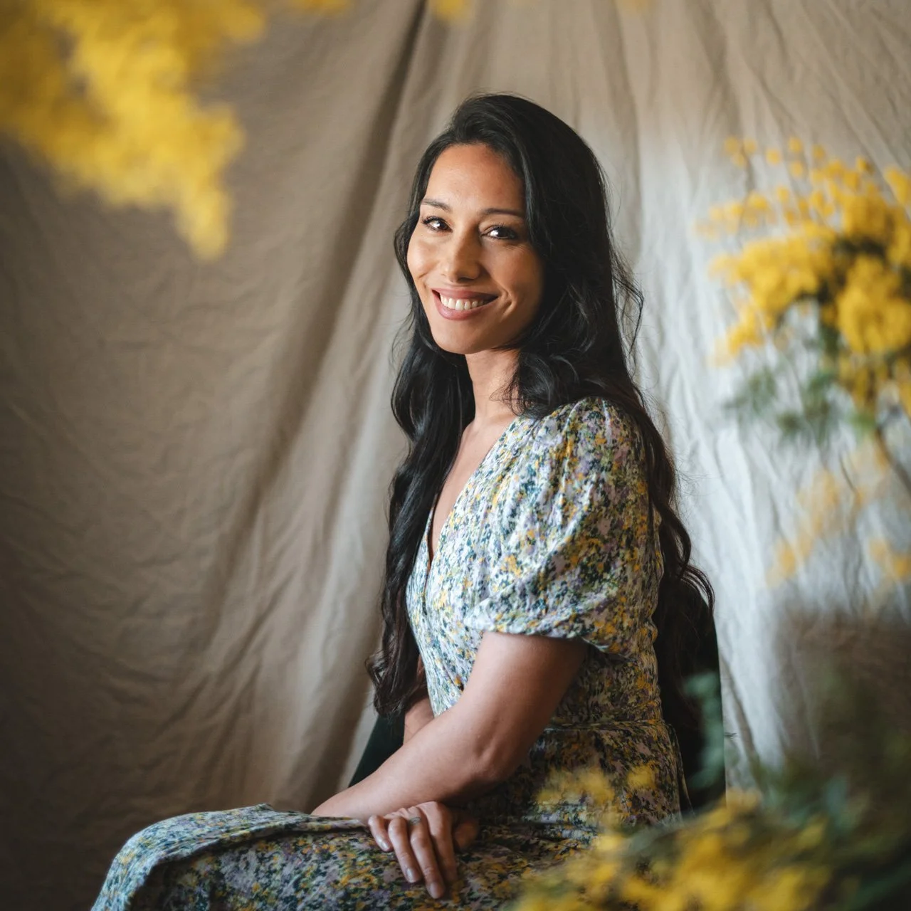 A woman with long dark wavy hair and a floral dress, smiling while sitting indoors with yellow flowers and beige fabric in the background.