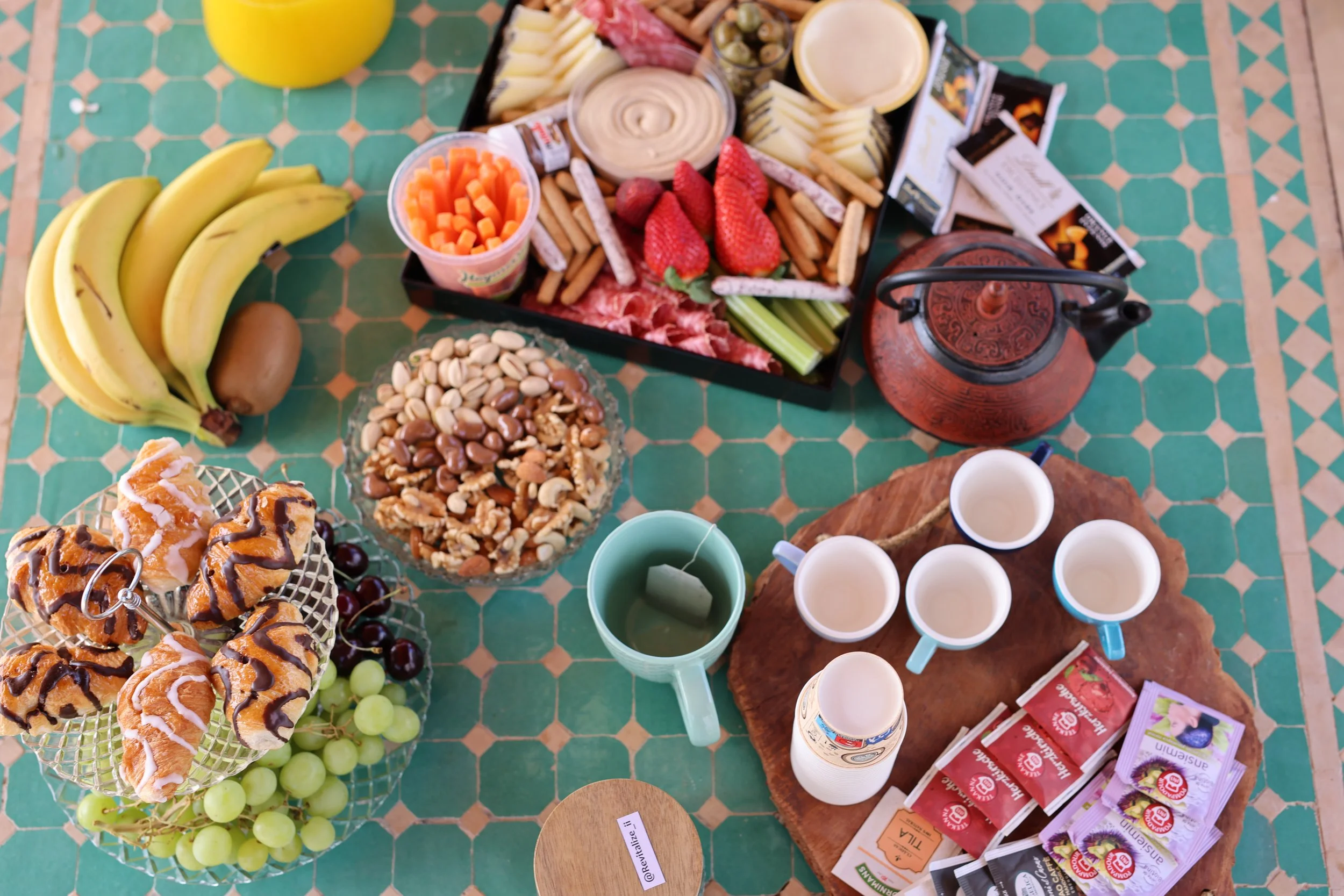 A breakfast spread featuring bananas and a kiwi, a bowl of mixed nuts, a plate of croissants with chocolate drizzle and grapes, a platter with strawberries, cheese, crackers, and sliced cucumber, a teapot, four cups with a hot beverage, and assorted 