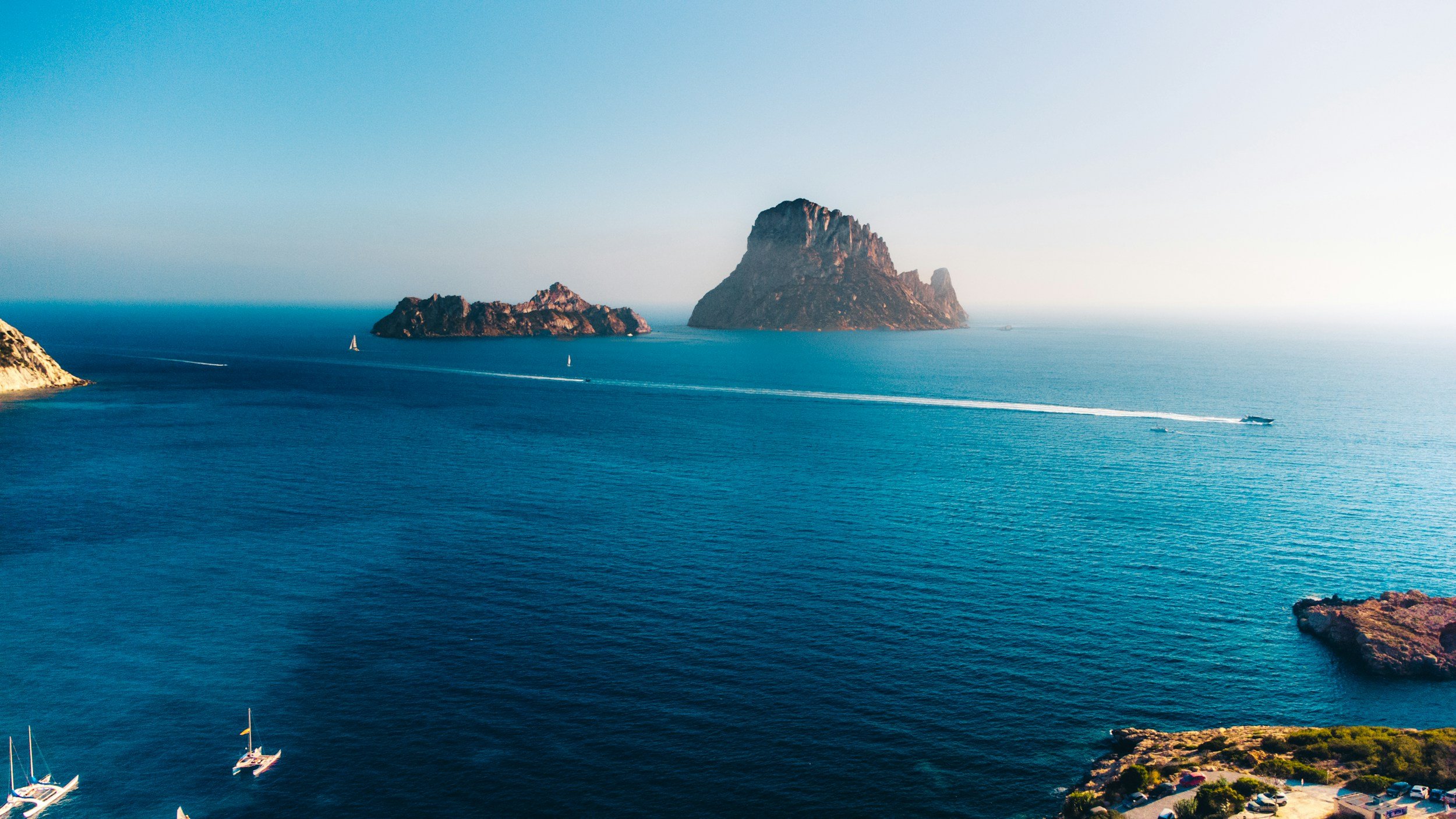 Aerial view of the sea with several small sailboats and a speedboat leaving a wake, rocky islands in the distance, and a coastline with buildings and greenery in the foreground.