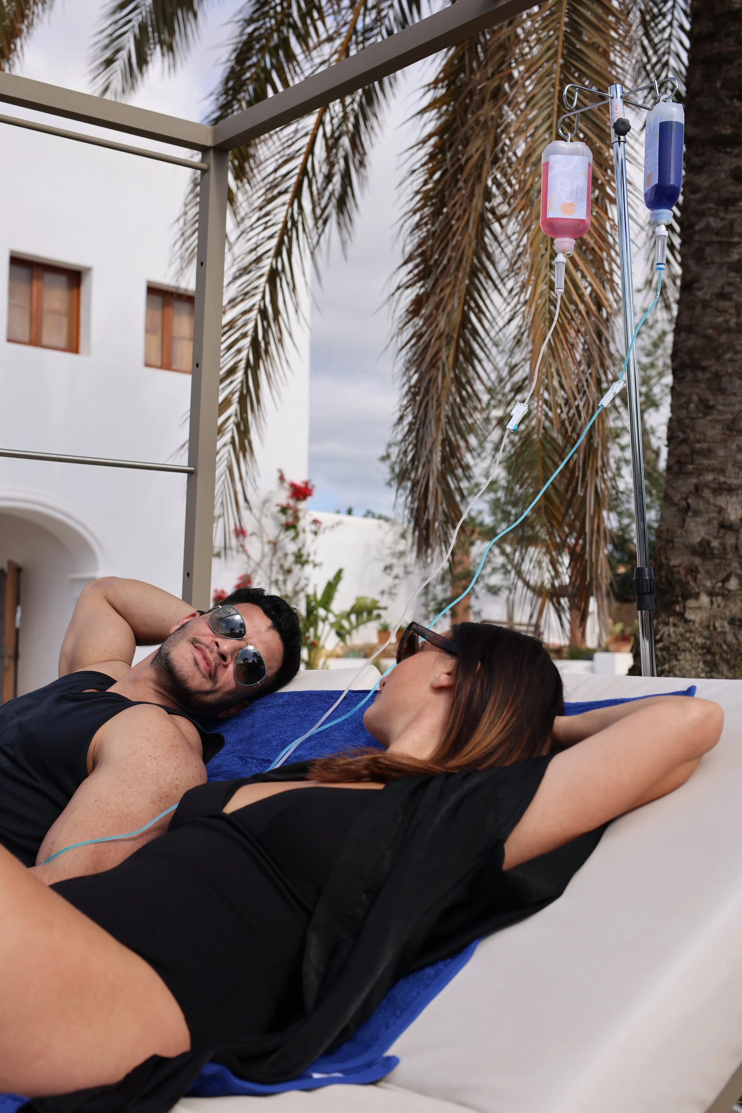 A man and woman relaxing on a lounge chair outdoors with IV drips beside them, surrounded by palm trees and a white building.
