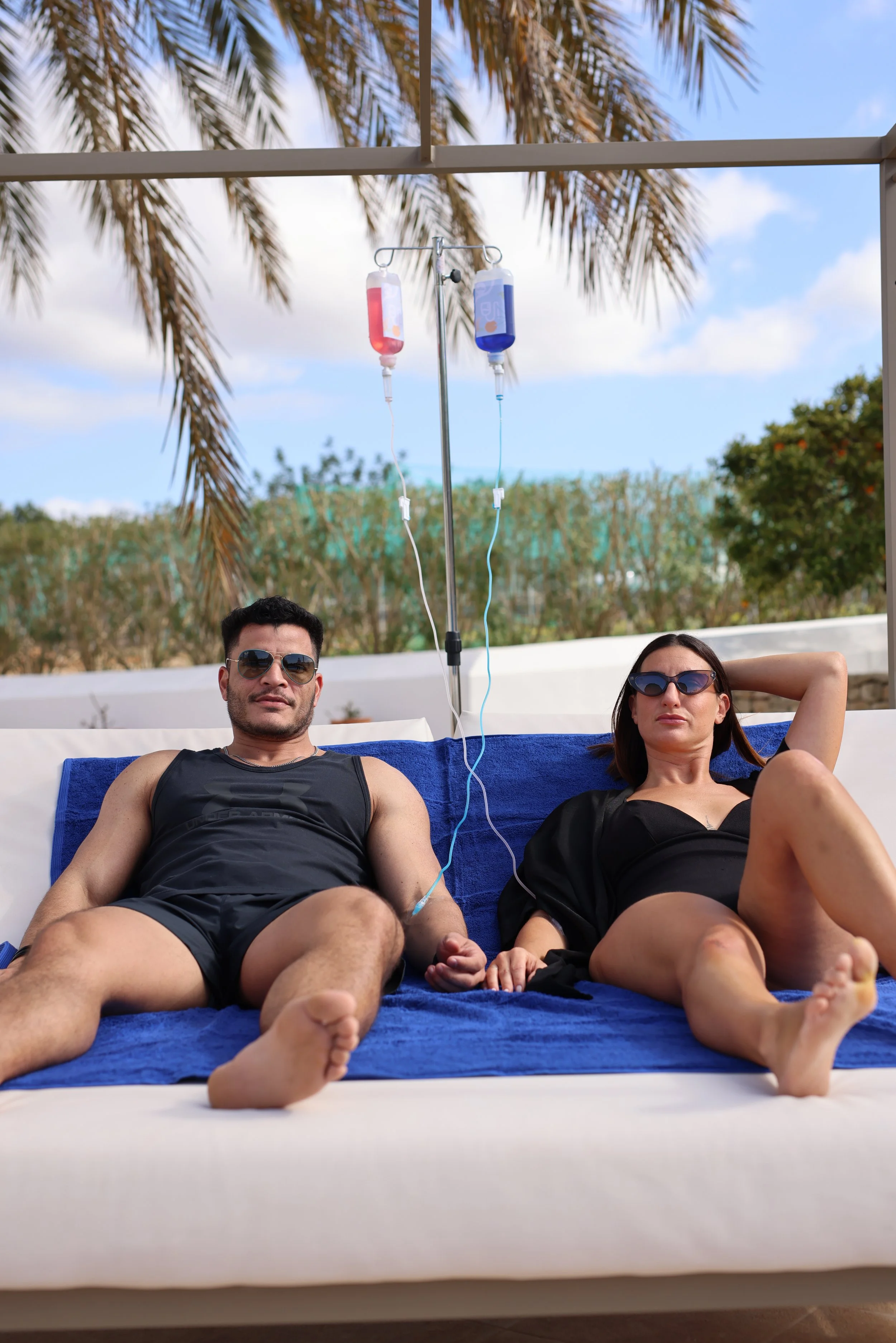 A man and woman lying on a beach lounge chair with IV bags hanging above them, outdoors with palm trees and blue sky in the background.