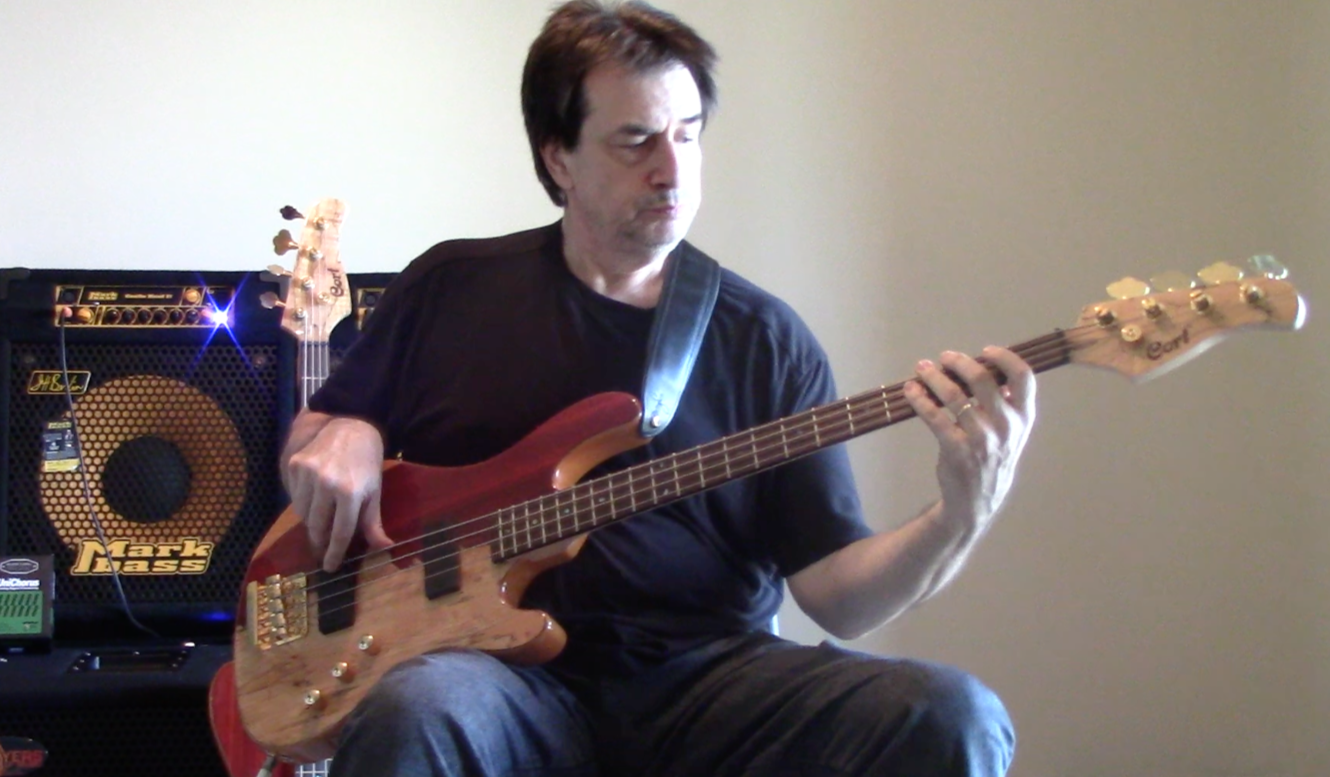 A man with dark hair playing a red and natural wood electric bass guitar, sitting, with a small amplifier in the background.