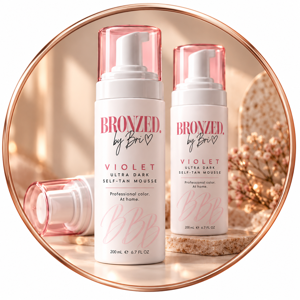 Two bottles of BRONZED by Bri Violet Ultra Dark Self-Tan Mousse with pink caps, one lying down and one upright, on a marble surface with a pink flower arrangement and a decorative stone backdrop.