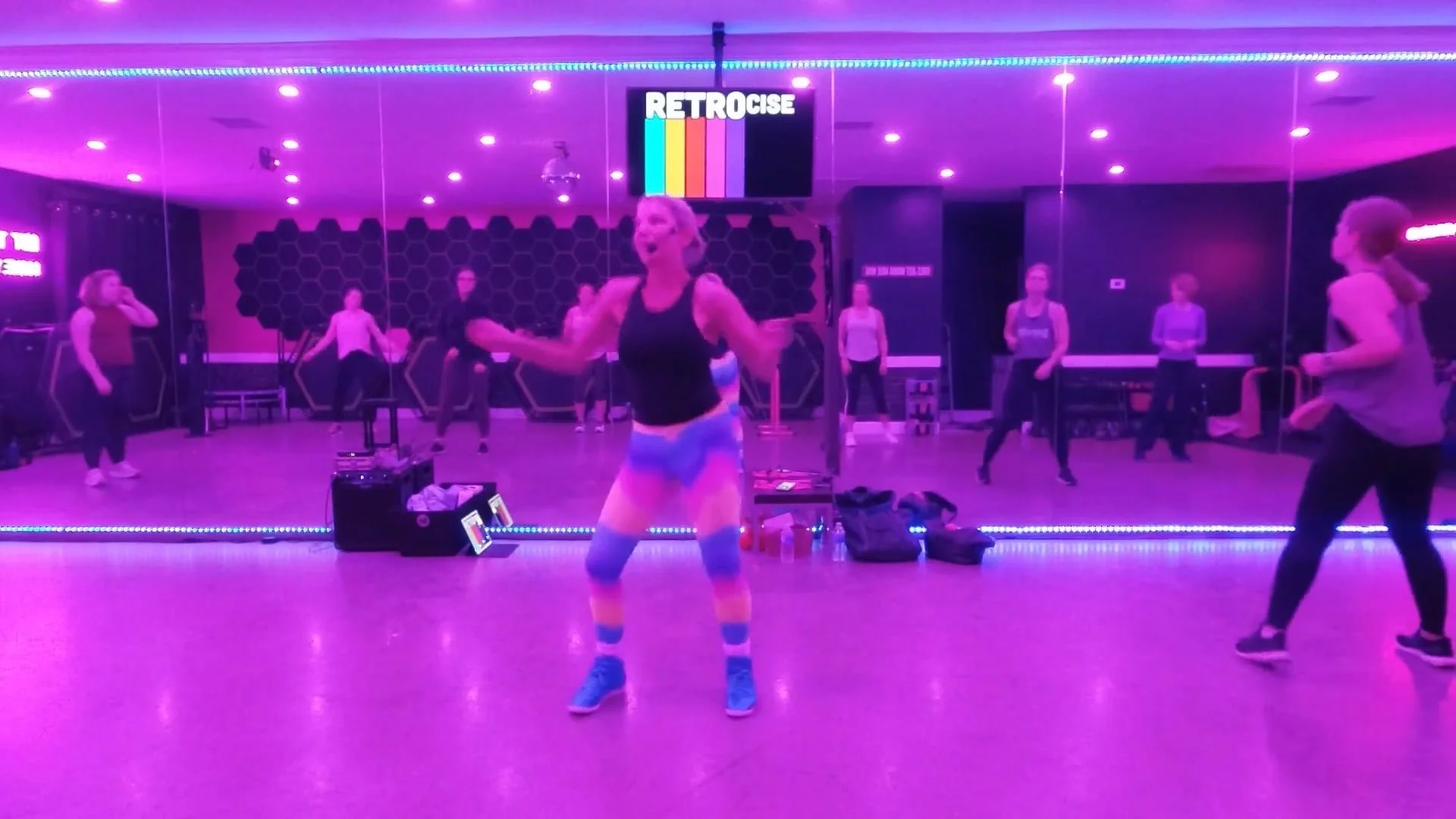 A woman exercising on a small trampoline in a room with black walls, blue speakers, and colorful lighting. She is lying on her back on the trampoline, pointing upward with one arm and the other arm extended to her side. A green exercise ball is on the floor nearby.