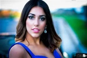 A woman with dark brown, shoulder-length hair and makeup, wearing a royal blue dress, standing outdoors with a blurred background of greenery and a pathway.