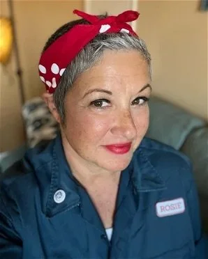 A woman with short gray hair wearing a red polka dot headband and a blue work uniform with a name tag labeled "ROSIE."