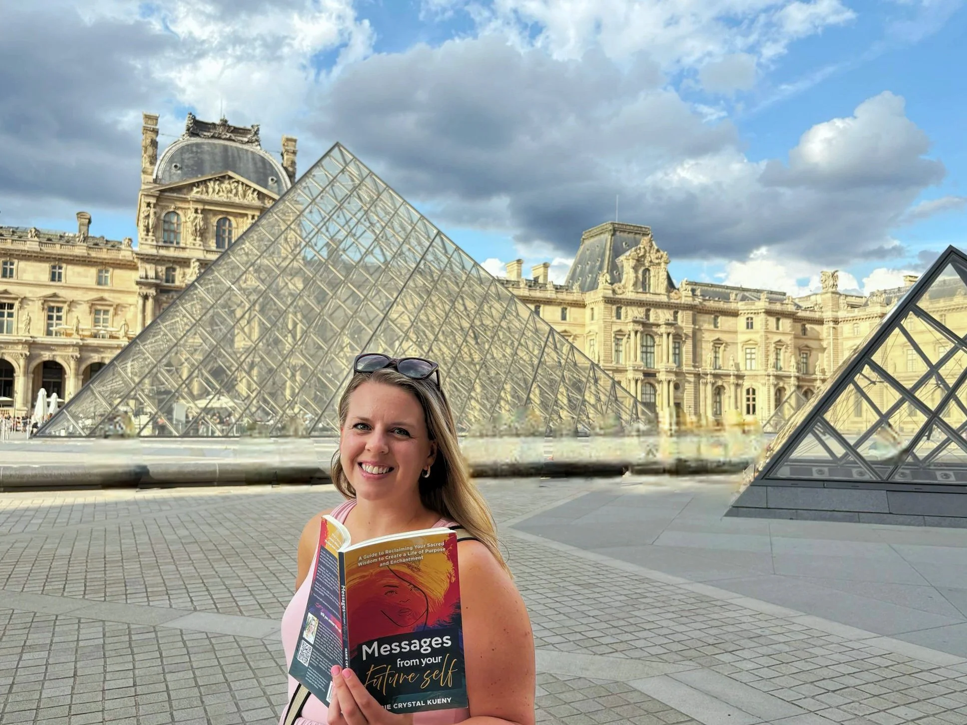 A woman holding a book titled 'Messages from your future self' in front of the Louvre Museum in Paris, France, with its glass pyramid and historic architecture under a partly cloudy sky.