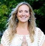 A smiling woman with long blonde hair wearing a white top, standing outdoors with green foliage in the background.