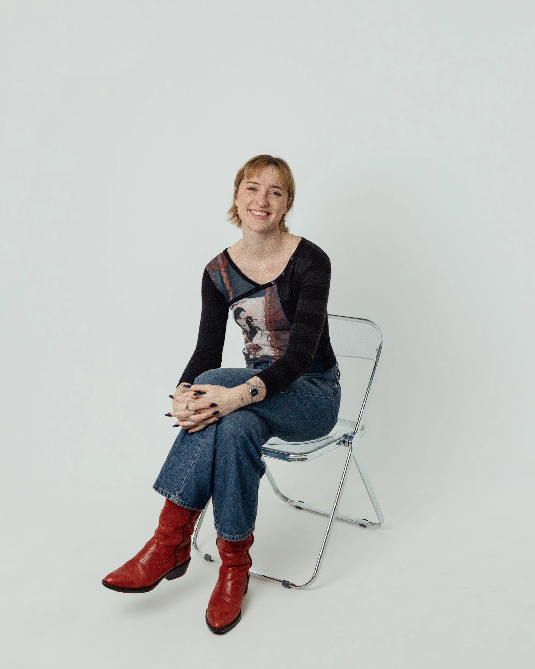 A young woman with blonde hair, smiling, seated on a modern metal chair against a plain white background. She is wearing a multicolored top, blue jeans, and red boots.