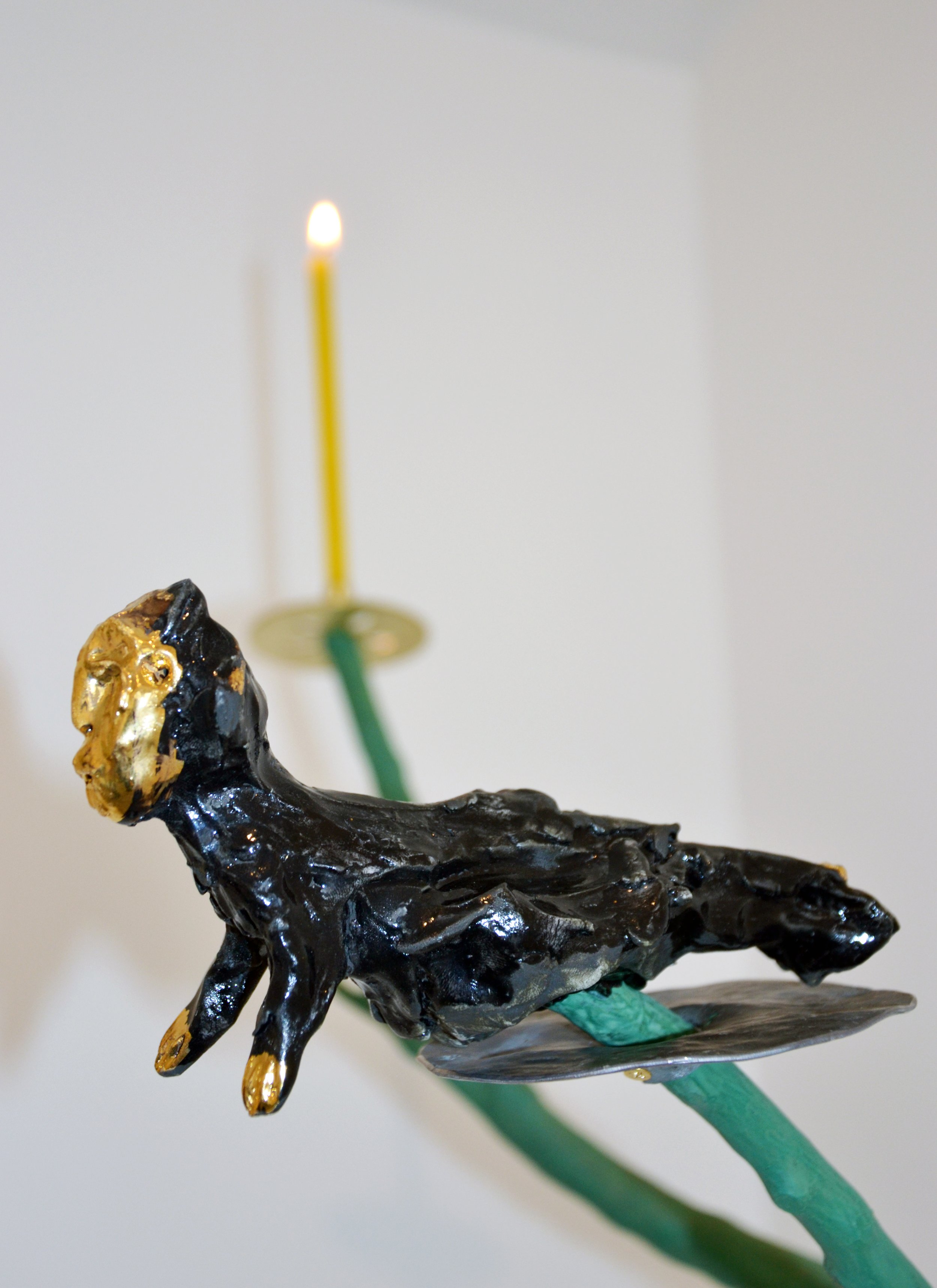 A black sculpture of a lion with gold accents on its face, supporting a green platform with a yellow candle on top, and a white background.