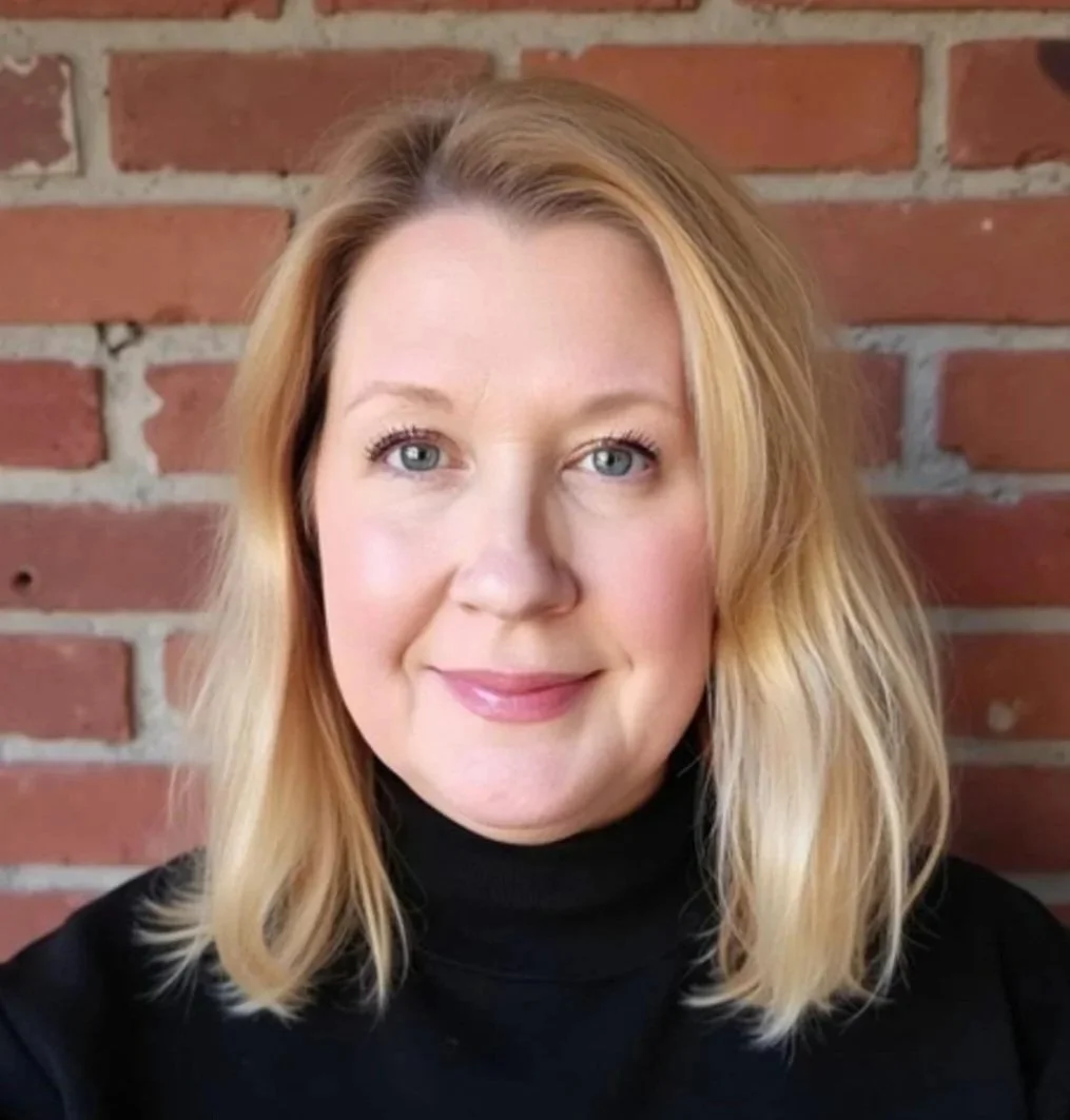 A woman with blonde hair wearing a black turtleneck standing in front of a red brick wall.
