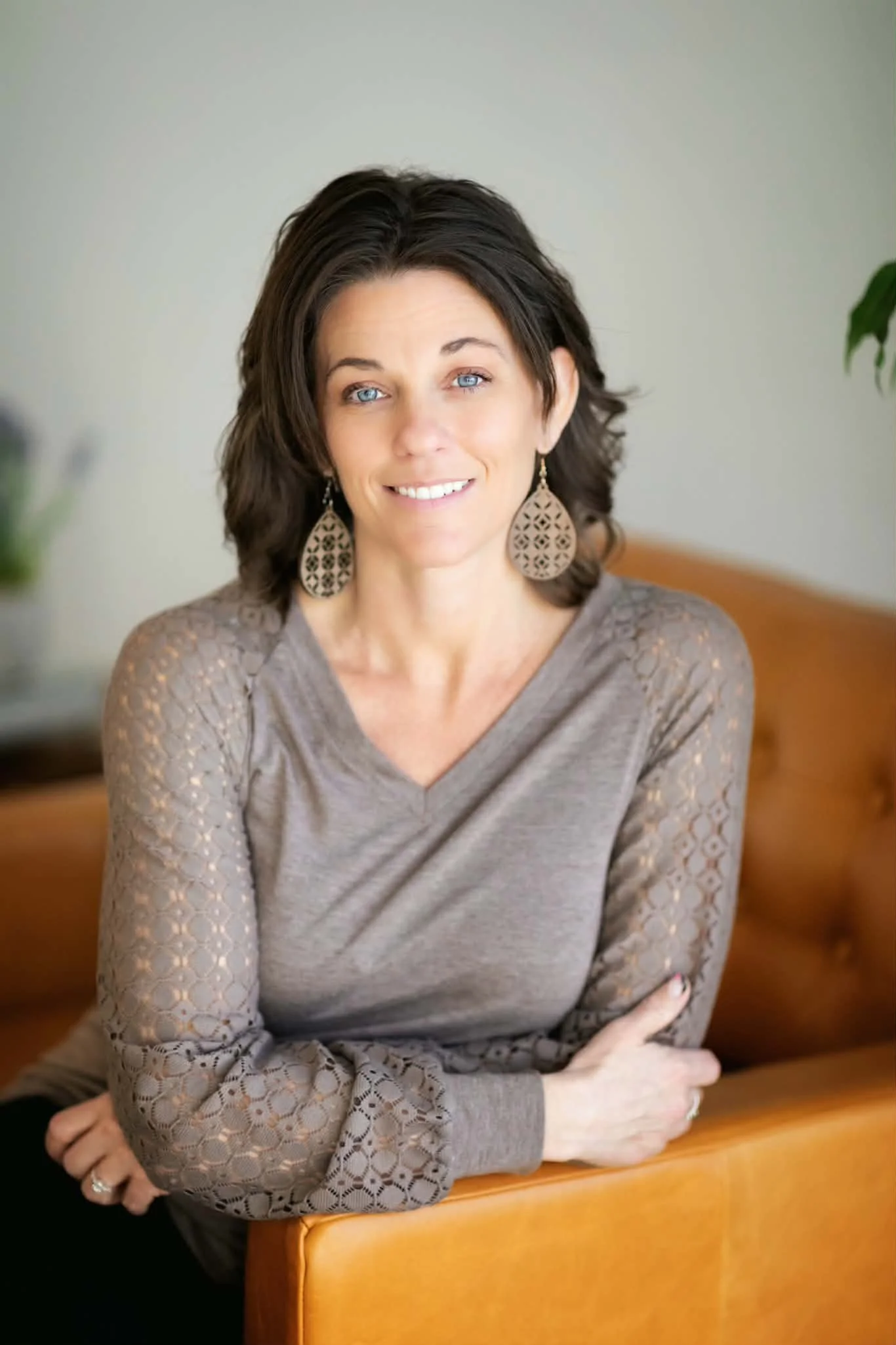 A woman with blue eyes and dark brown hair, smiling and sitting at a wooden table. She is wearing large, decorative earrings and a long-sleeved, beige top with a pattern of holes on the sleeves. Her left hand is resting on the table, and her right arm is crossed over her body. The background is plain with a muted green wall and a blurred plant to the right.