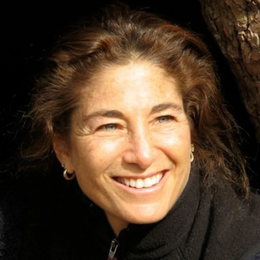 Close-up of a smiling woman with brown hair, blue eyes, and earrings, outdoors near a tree bark background.