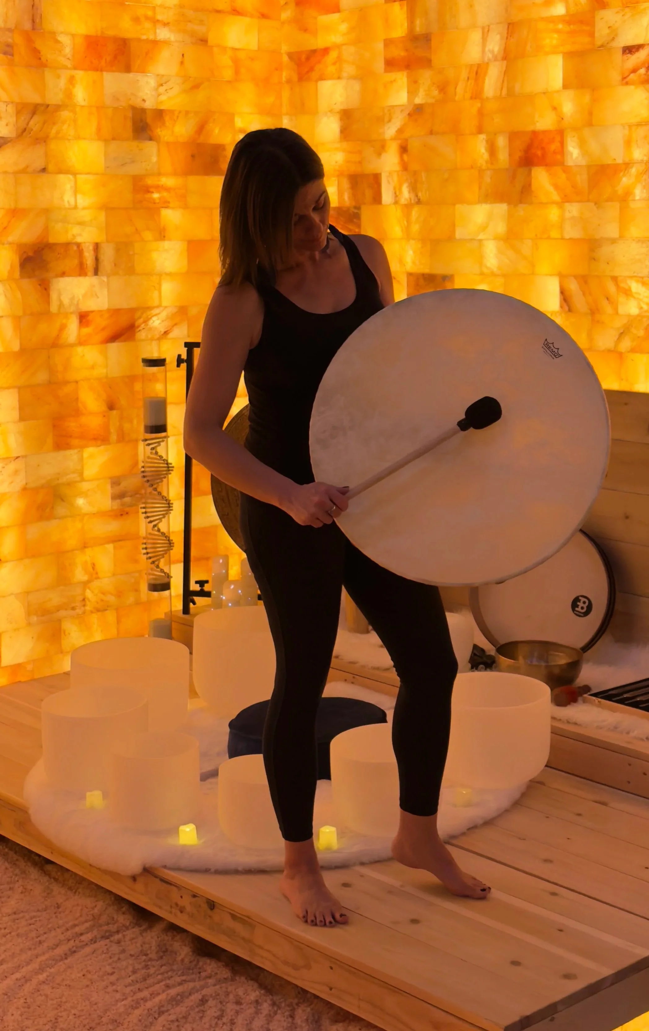 A woman standing on a wooden platform, holding a large drum with a mallet, surrounded by glowing candles and Himalayan salt lamps, in a cozy-lit room with salt wall decor.