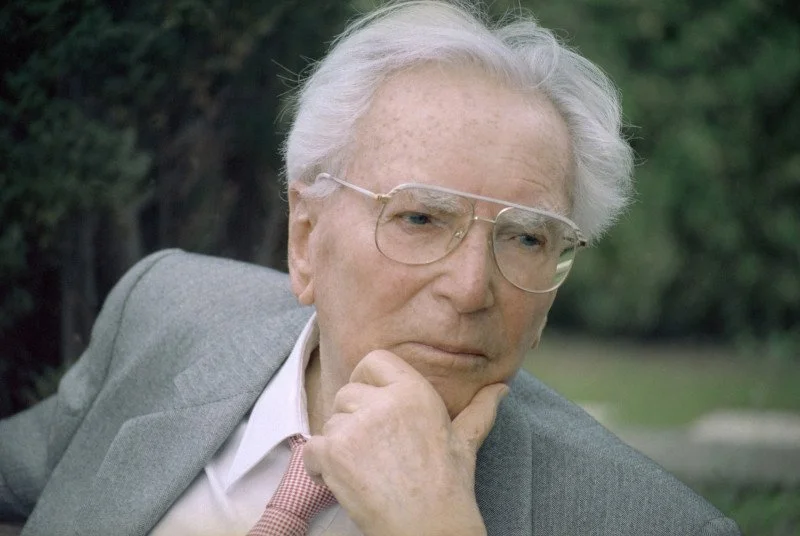 Old man with gray hair and glasses in a gray suit, sitting outdoors with a thoughtful expression, hand on chin.
