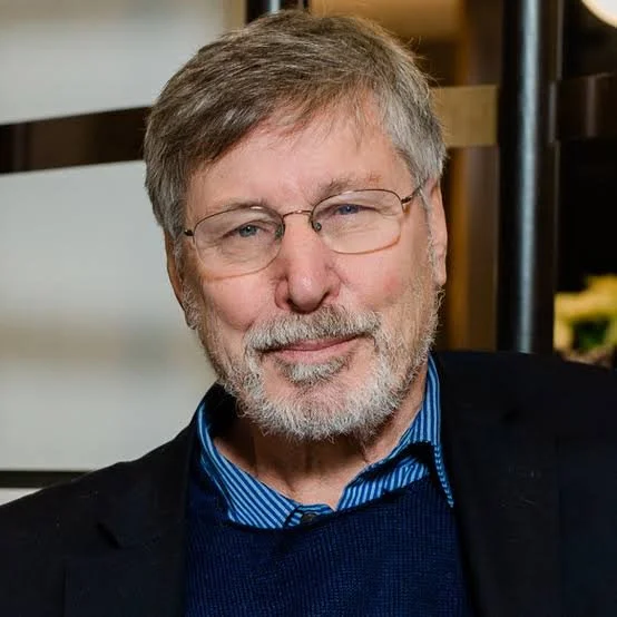 An older man with glasses, gray hair, and a beard, wearing a blue shirt and dark blazer, smiling at the camera indoors.