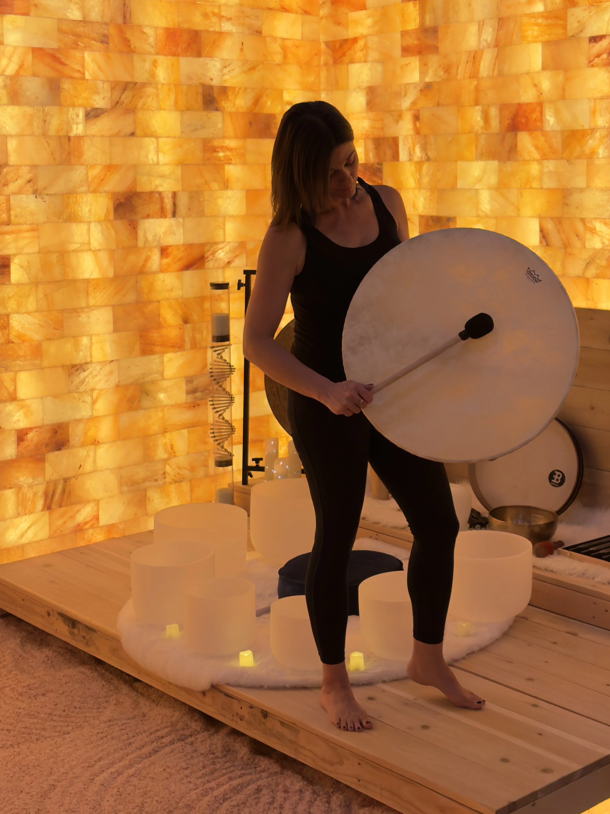 Woman standing on a wooden platform holding a large drum with a mallet, surrounded by candle-like lights and Himalayan salt lamps, in a softly lit room.