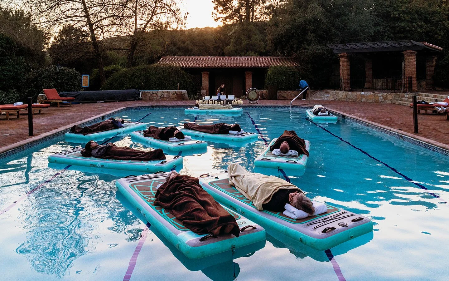 People receiving a floatation therapy session in an outdoor pool, lying on floating beds with heads resting on pillows, at sunset.