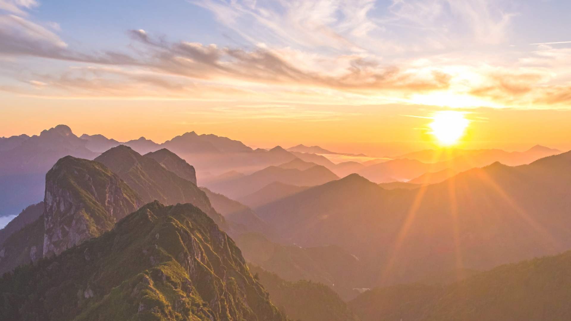 Sunrise over mountain range with partly cloudy sky and layered mountains in the background.