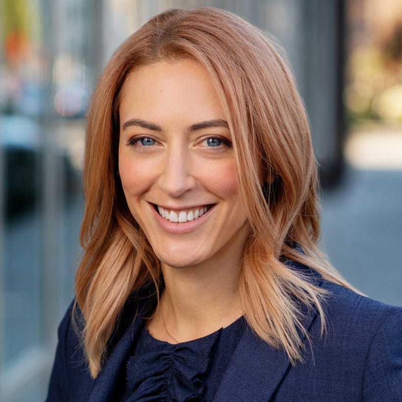 A woman with shoulder-length red hair and blue eyes smiling outdoors in a business suit.