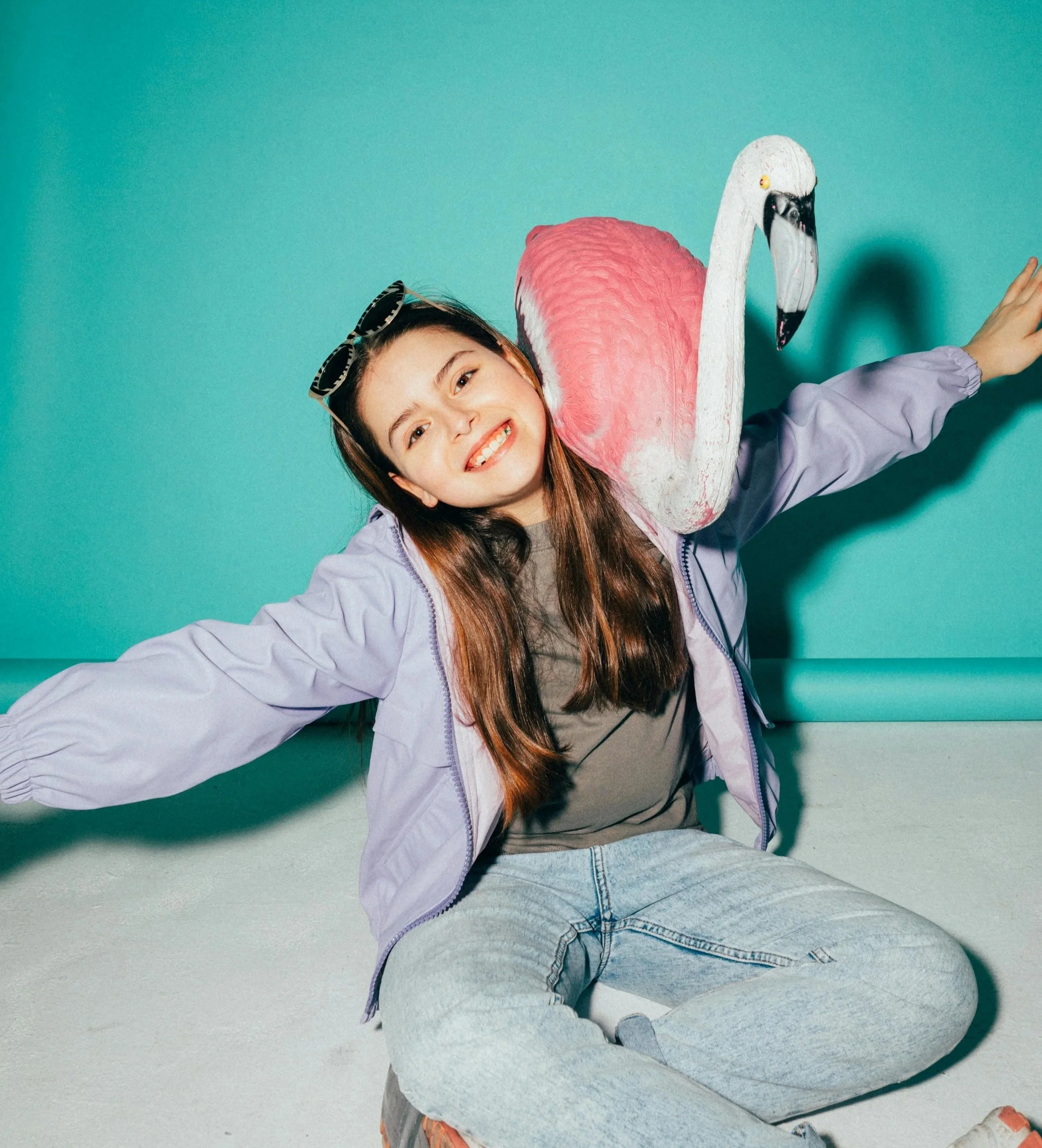 A girl smiling and sitting cross-legged on the floor with an open arm pose, wearing sunglasses on her head, a lavender jacket, a gray T-shirt, light blue jeans, and a pink flamingo-shaped backpack.