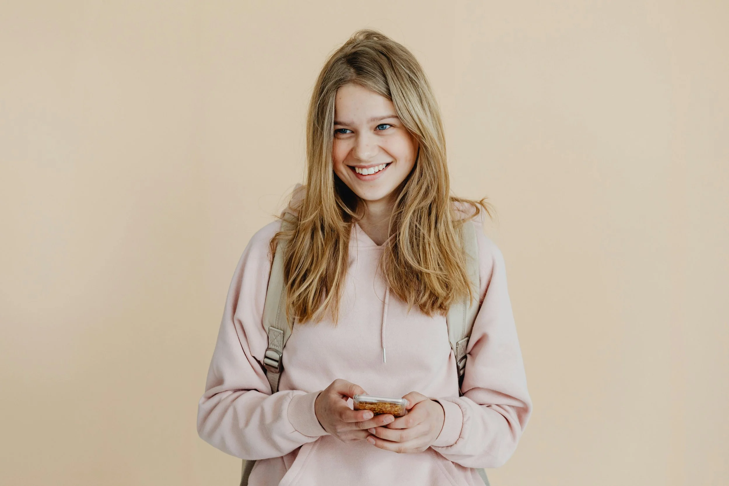 A young woman with long blonde hair smiling while looking at her smartphone, wearing a light pink hoodie and a backpack, standing against a plain beige background.