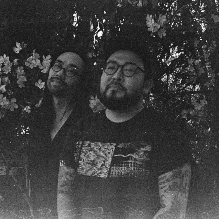 Two men with glasses standing outdoors in front of a flowering bush, one with a beard and the other with long hair, both facing camera.