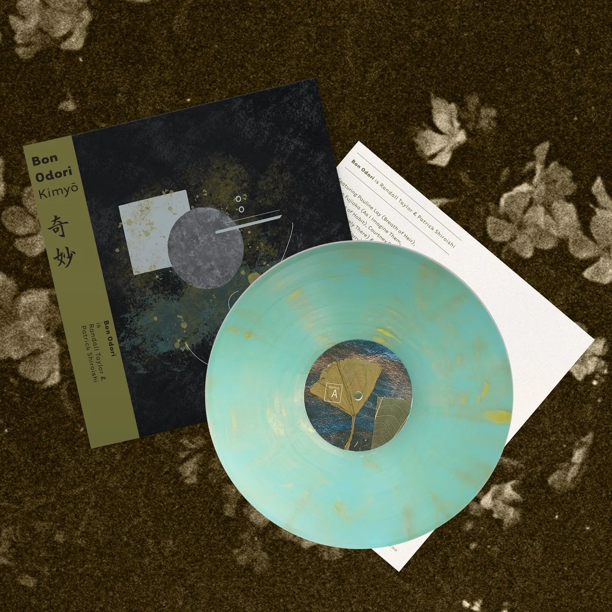 A vinyl record with a light green and yellow marbled pattern, placed on a white paper, along with a black and gold album cover featuring abstract artwork, and a white sheet of paper with printed text, all on a dark surface with scattered white flower petals.