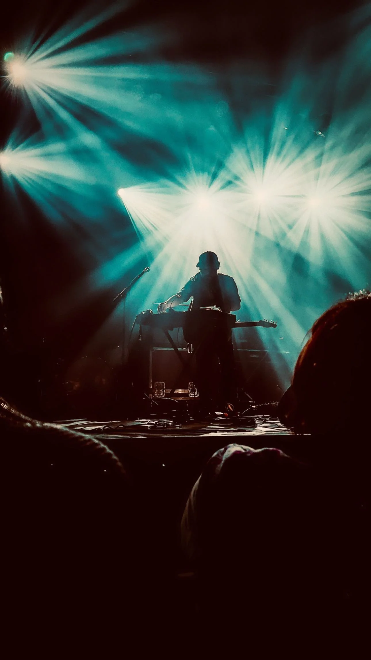 A musician performing on stage with bright teal and white lights shining behind him, silhouetted against the stage lights, with audience members visible in the foreground.