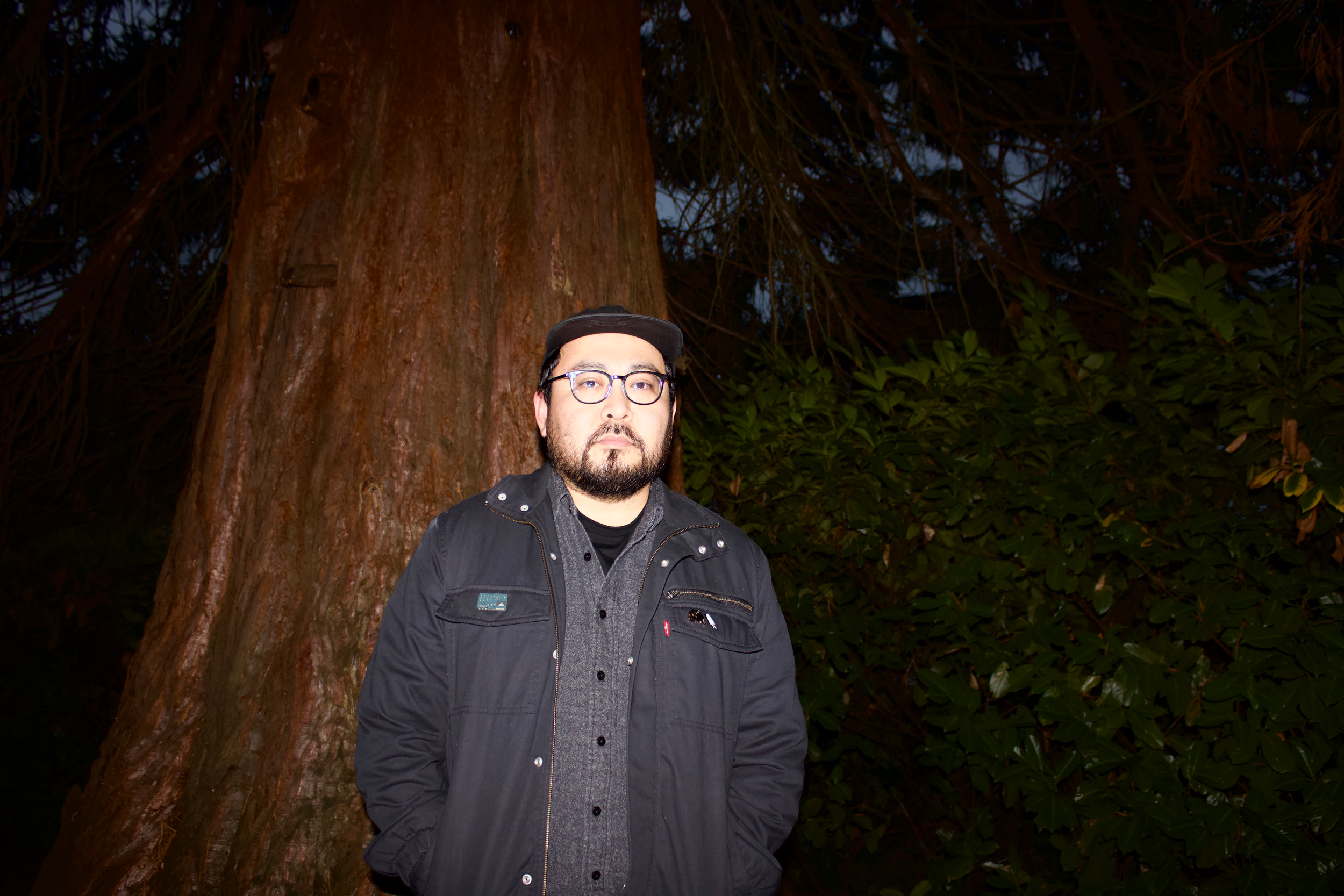 A man wearing glasses, a black jacket, and a cap standing in front of a large tree with dense foliage at night.