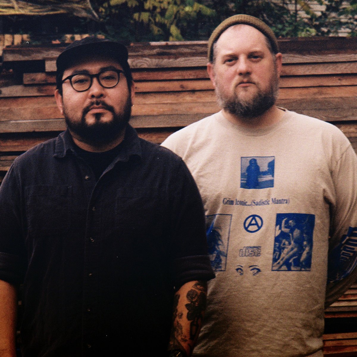 Two men standing outdoors in front of a wooden fence. The man on the left has glasses, facial hair, and tattoos on his arm. He is wearing a black cap and dark shirt. The man on the right has a beard, is wearing a tan shirt with blue graphics, and a tan beanie.