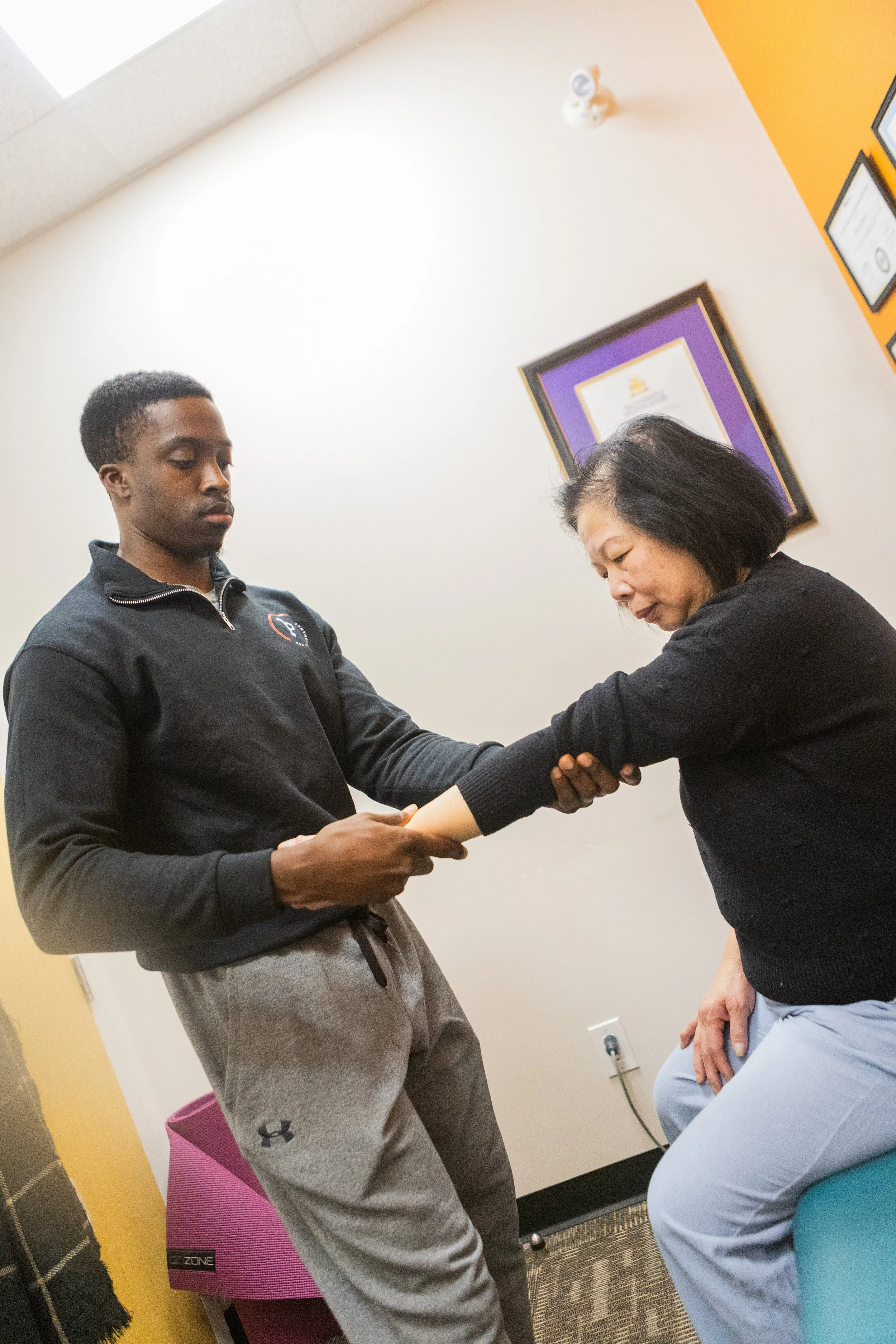 A young man helps an older woman stretch her arm in a therapy session, seated in a room decorated with framed certificates and colorful walls.