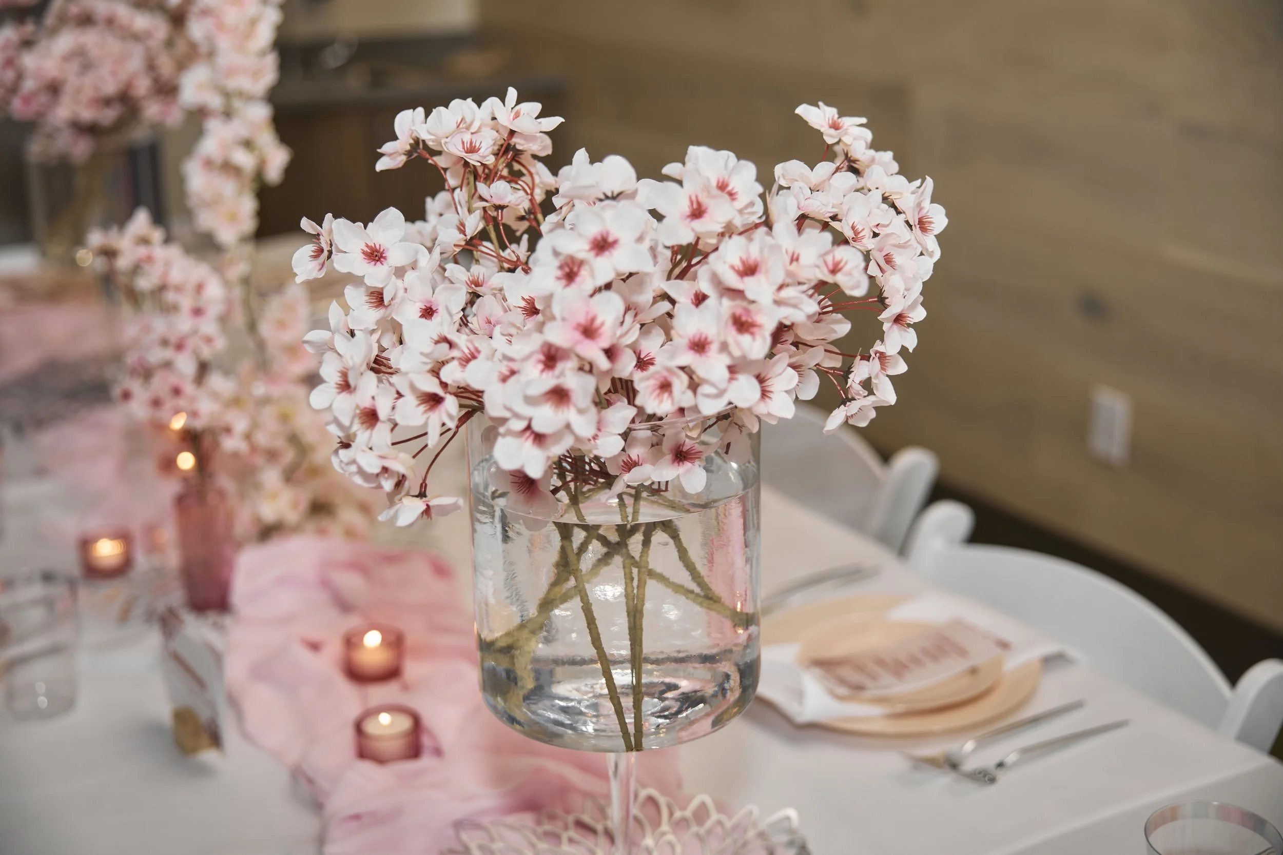 A tall glass vase filled with water and pink cherry blossom flowers as a centerpiece on a decorated event table, with pink candles and folded napkins in the background.