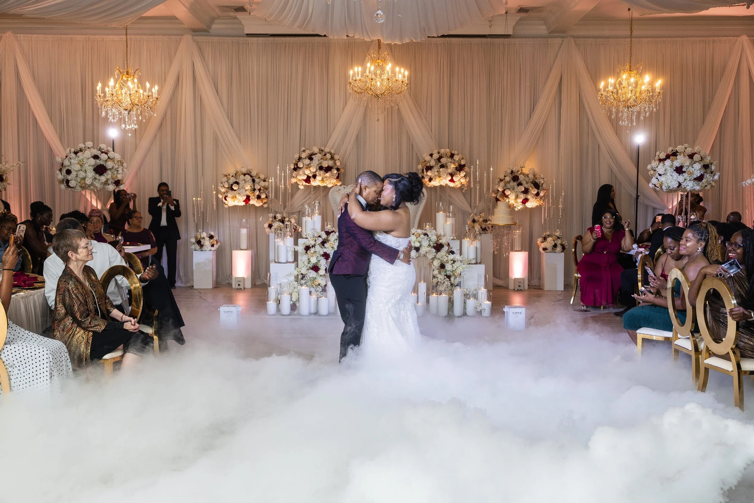 A bride and groom sharing a dance on a foggy dance floor during their wedding reception, surrounded by guests, with an elegant backdrop of white drapes, chandeliers, and floral arrangements.