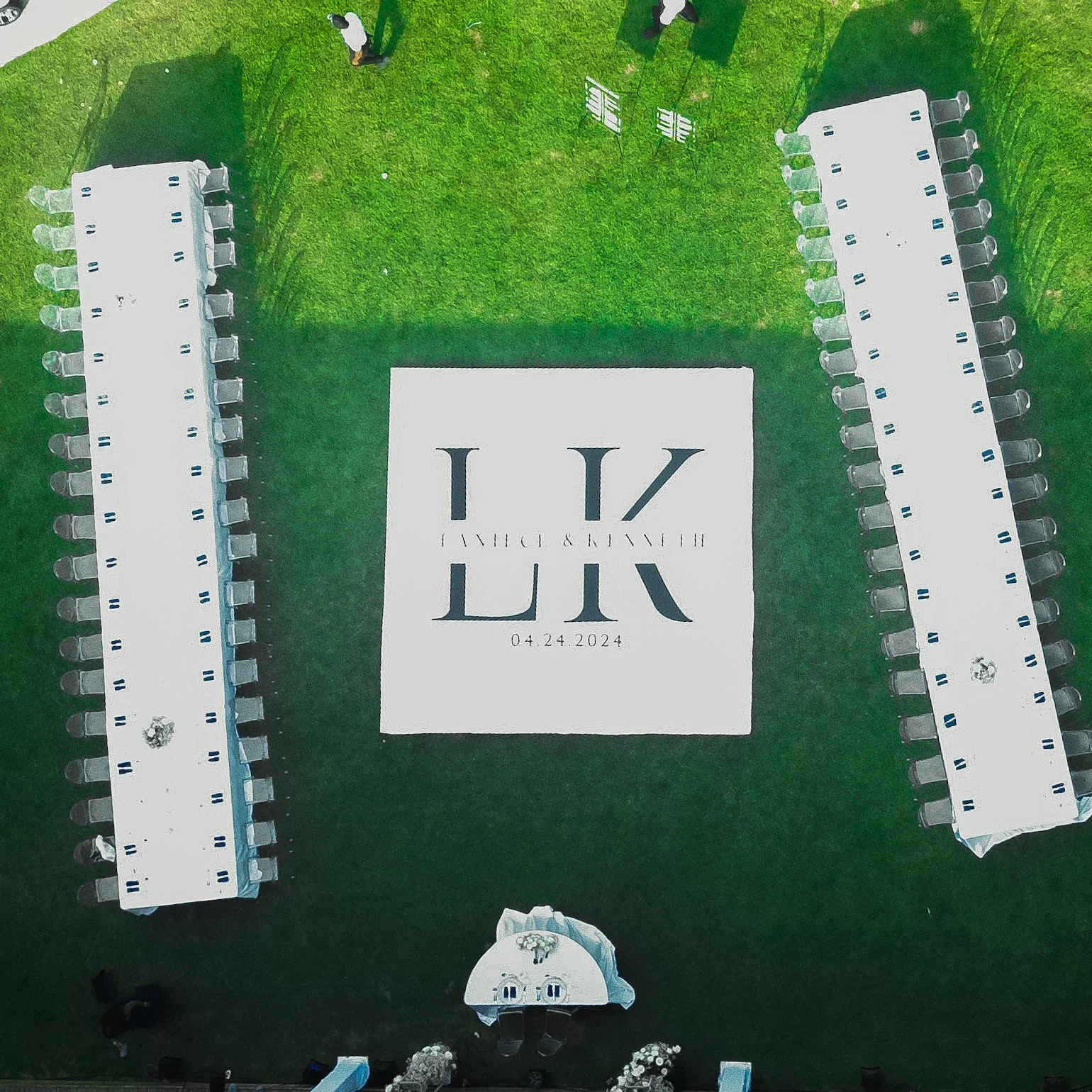 Aerial view of an event setup on grass with two long dining tables and a rug with initials "LK" and date 04.24.2024.