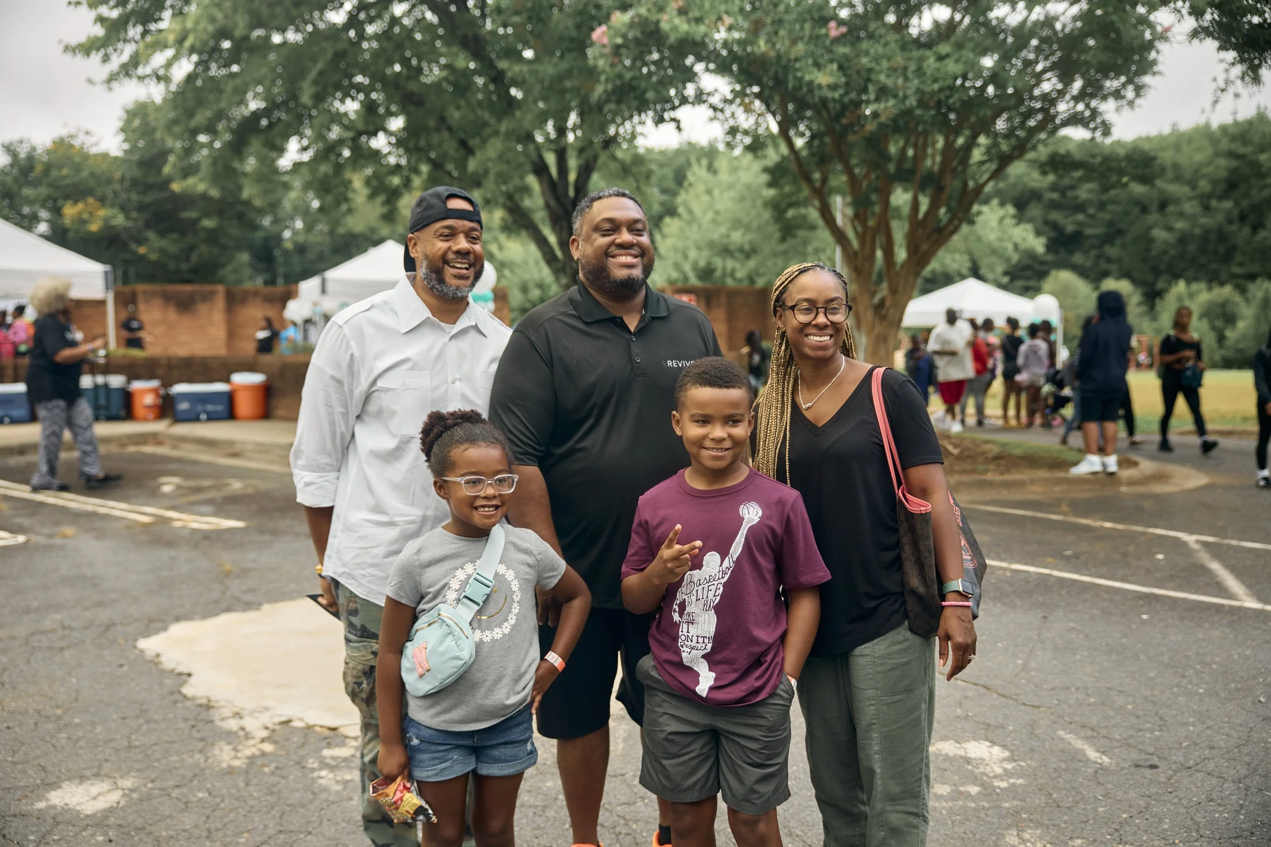 A group of five people, two kids and three adults, standing outdoors at a park or community gathering, smiling for the photo, with other people and tents in the background.