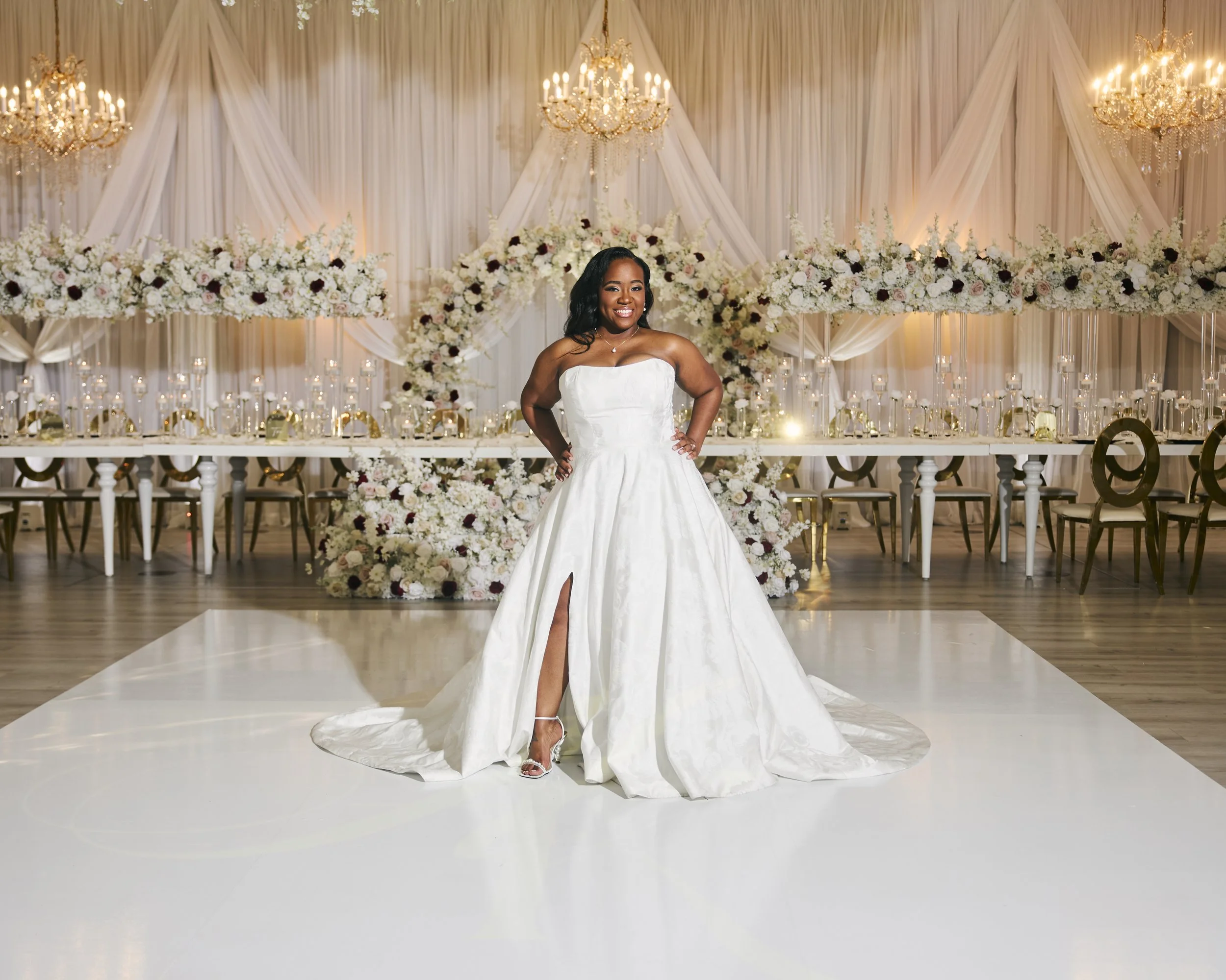 A woman in a white wedding dress standing in a decorated wedding venue with floral arrangements and chandeliers.