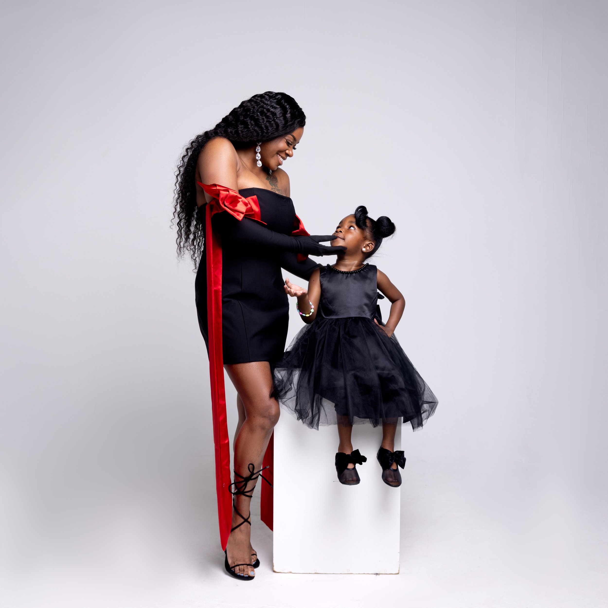 A woman in a black and red off-shoulder dress with long curly hair and black lace-up heels is smiling and holding the chin of a young girl sitting on a white pedestal. The girl, dressed in a black sleeveless dress with a tulle skirt and black shoes w