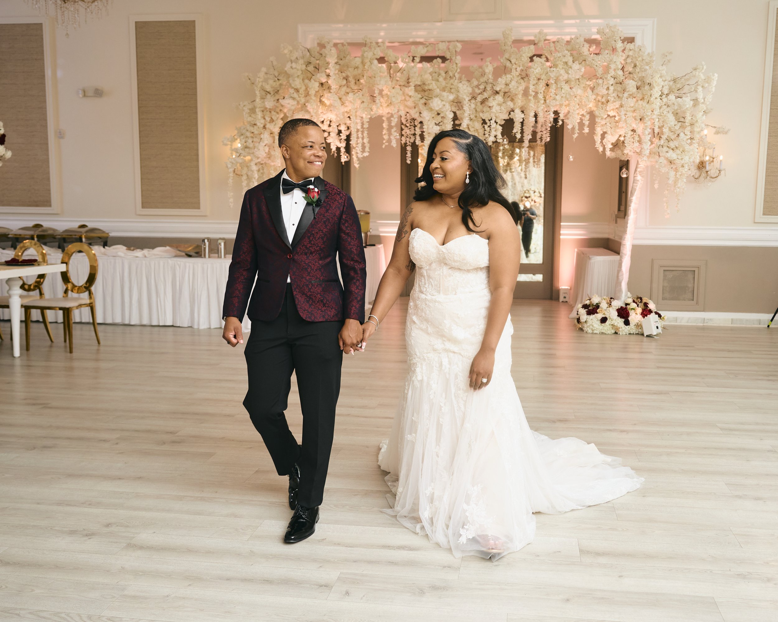 A couple dressed in wedding attire holding hands and smiling at each other on a dance floor, with a decorated floral arch in the background.
