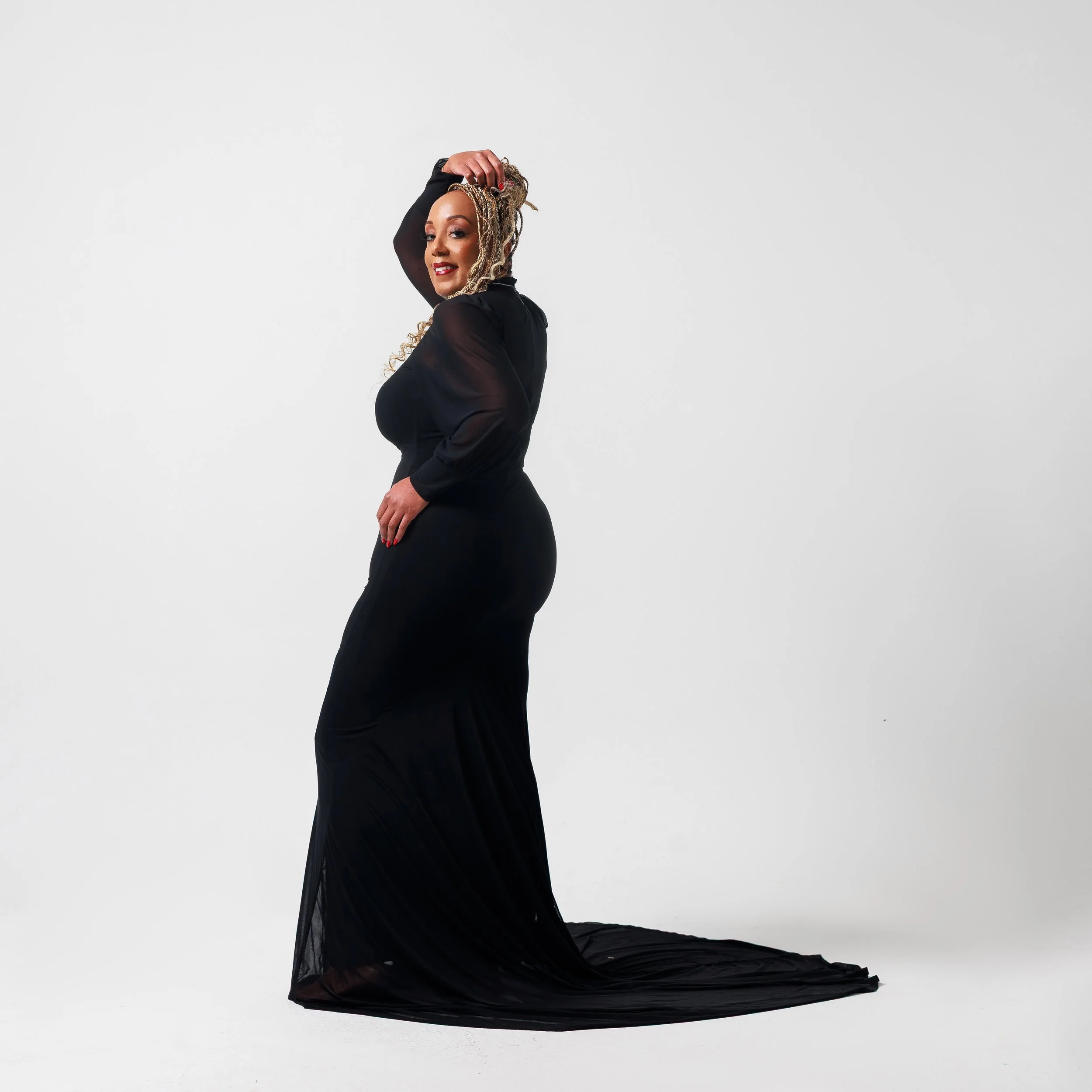 A woman with blonde curly hair in a black gown with sheer sleeves, posing against a plain white background.
