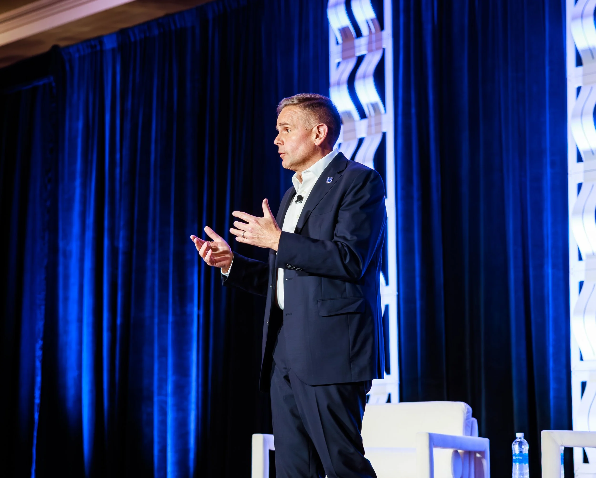 A man in a dark suit giving a presentation on a stage with blue curtains and abstract white decorative panels.