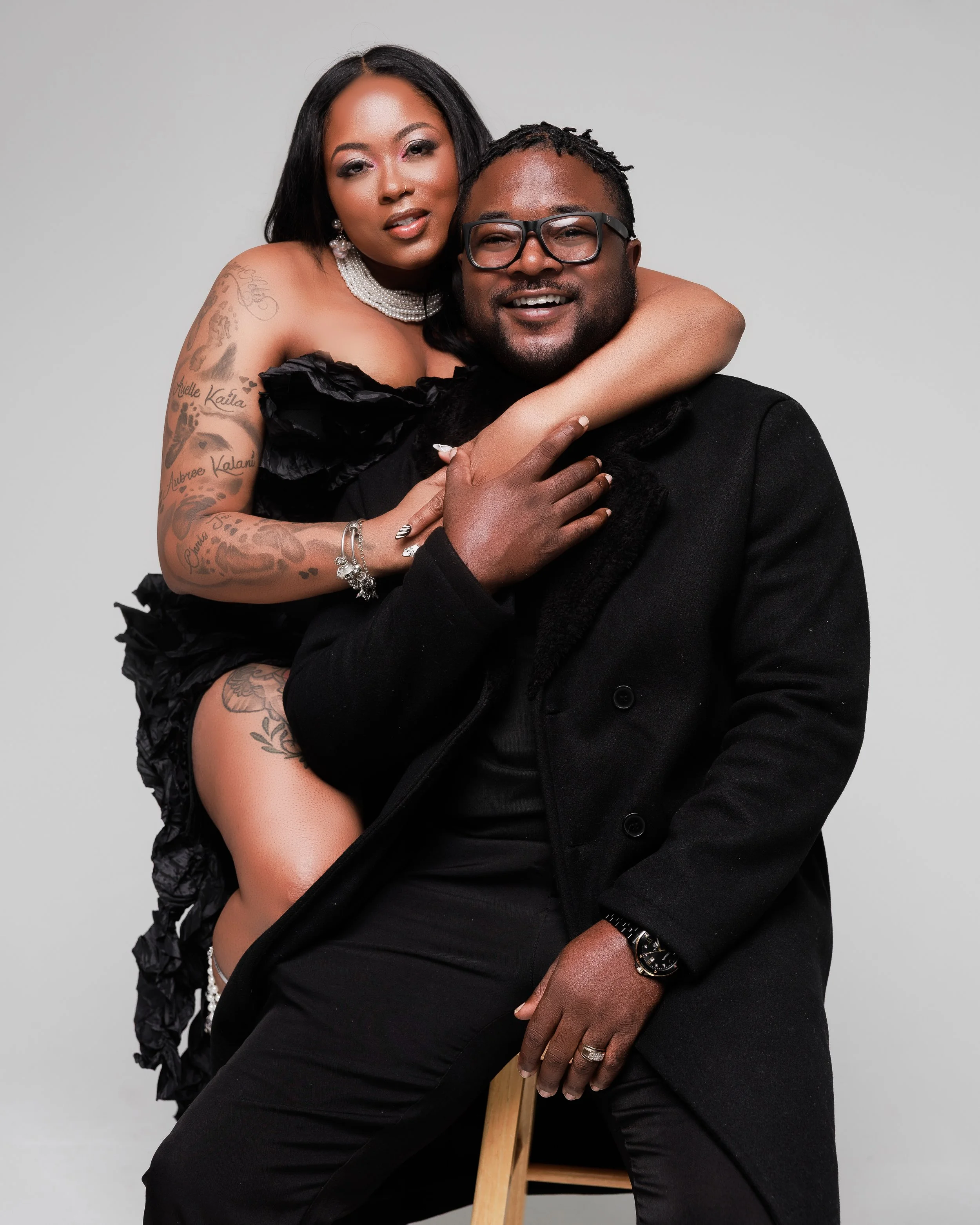 A smiling woman with long hair and tattoos on her arm sitting on a man's lap, with her arm around his neck, and the man sitting on a wooden stool, wearing glasses and a black coat. The woman is wearing a black dress and jewelry. They are in a studio with a plain background.