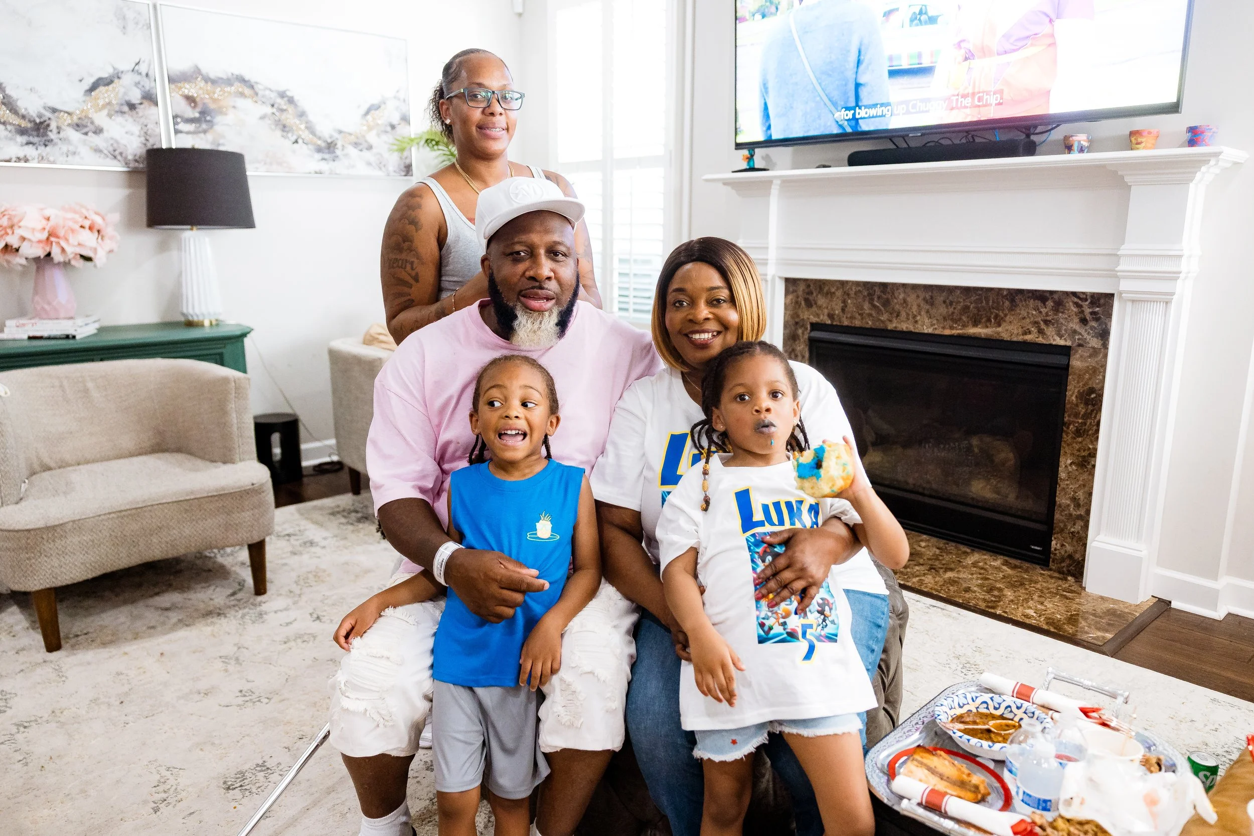 A family of six celebrating together in a living room, some wearing birthday-themed shirts, with a TV and fireplace in the background.