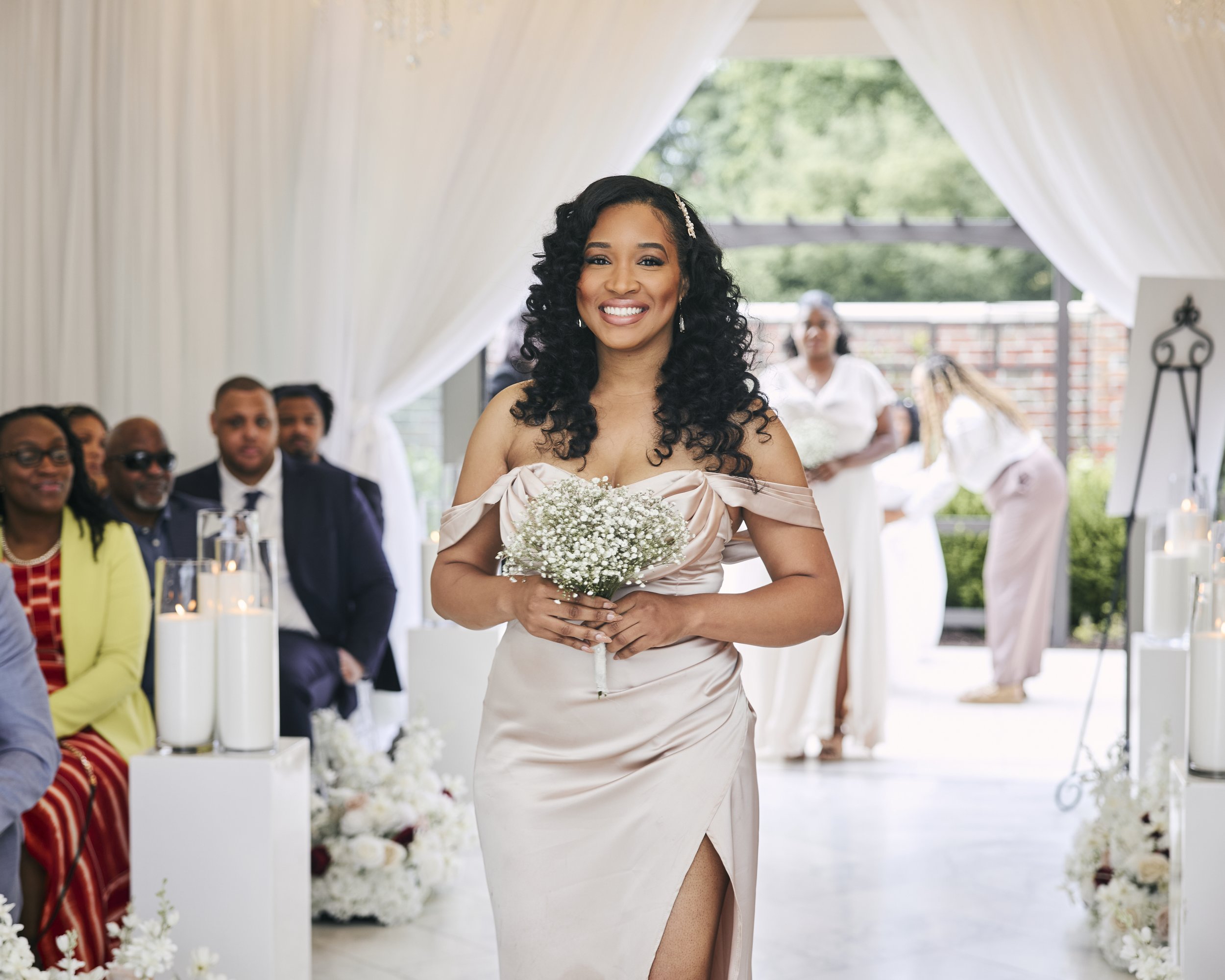 A woman in a silky, off-the-shoulder dress holding a small bouquet of white flowers, smiling at a celebration or wedding, with seated guests and assistants in the background under a white canopy decorated with candles and flowers.