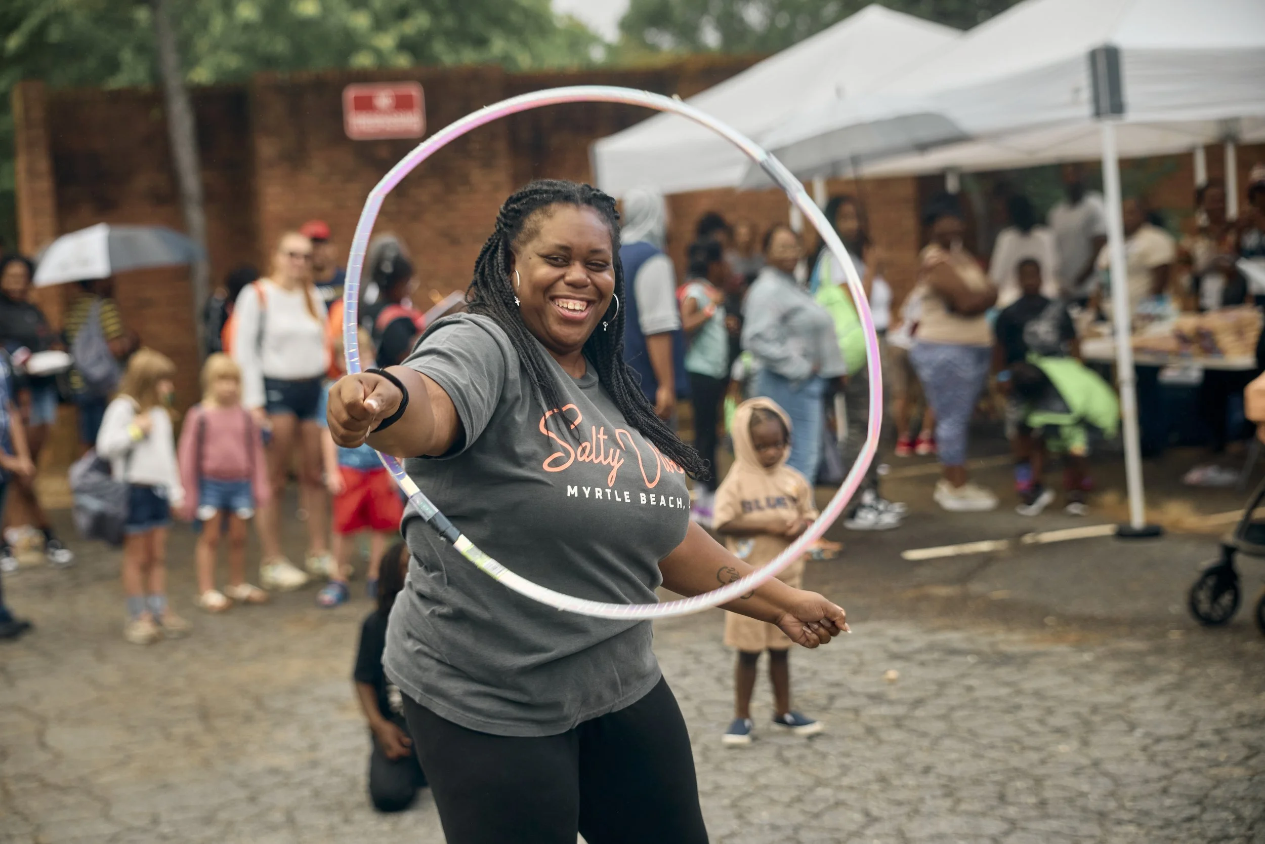 A woman smiling and playing with a hula hoop at an outdoor event with a crowd of people and tents in the background.